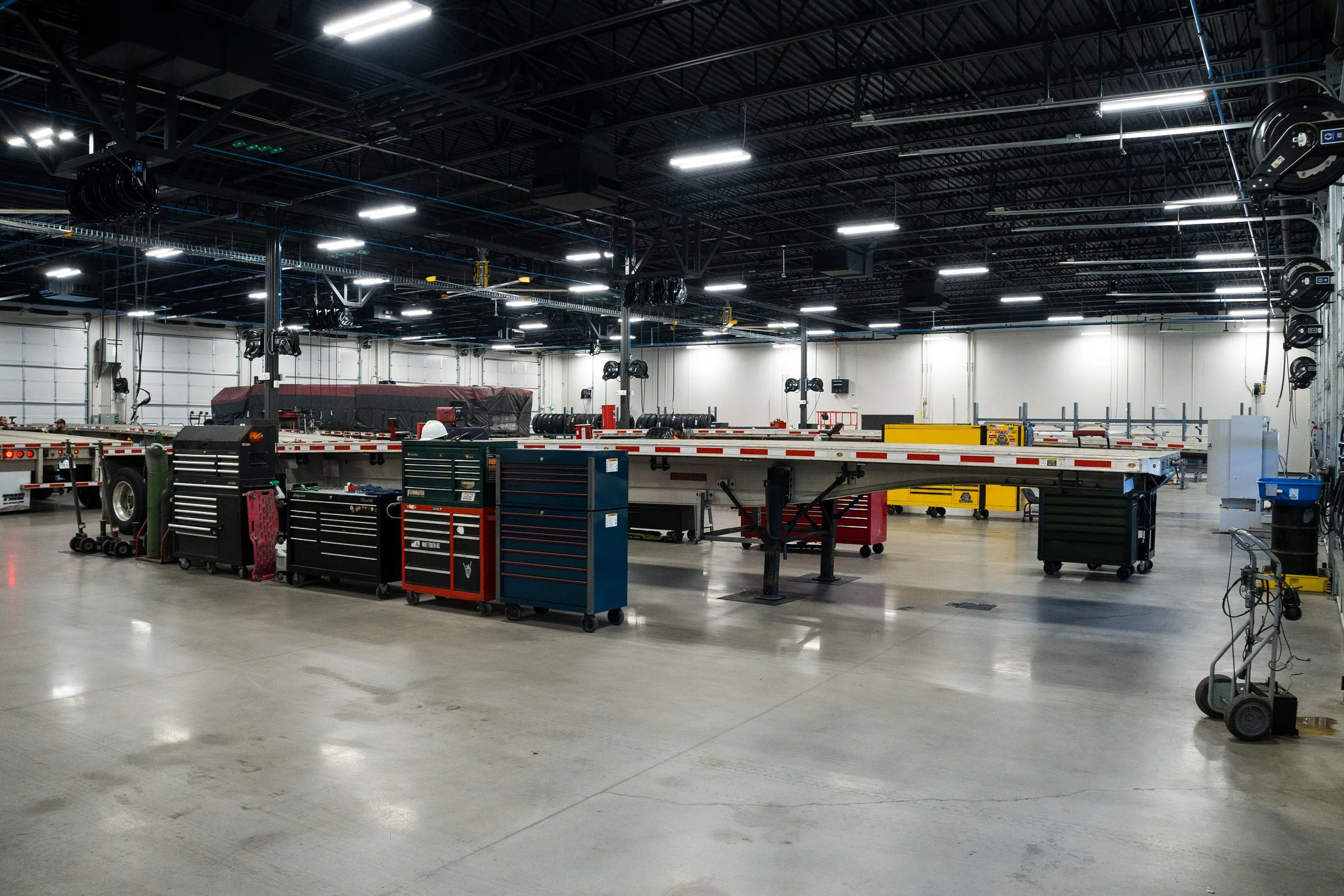 An indoor industrial or warehouse space with a high ceiling, overhead lighting, and various equipment and tools scattered across the floor. There is no visible merchandise or inventory.