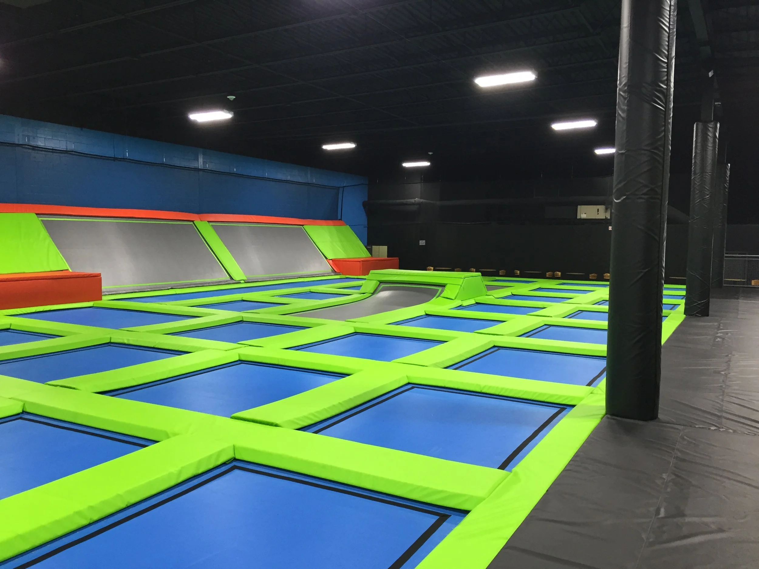 Indoor trampoline park with multiple trampolines with green borders, some small foam pits, black padded poles, and a dark ceiling with fluorescent lighting.