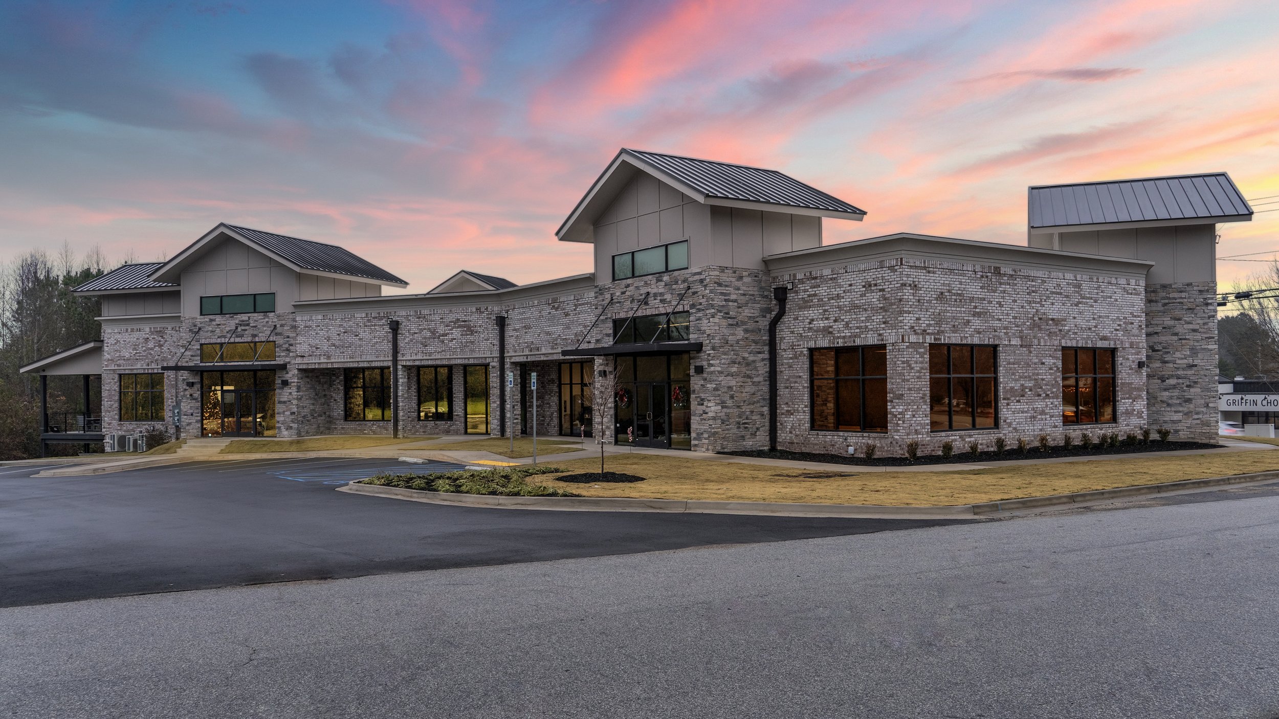 A modern commercial building with stone and metal exterior, large windows, and a small parking lot before it, during sunset with a colorful sky.