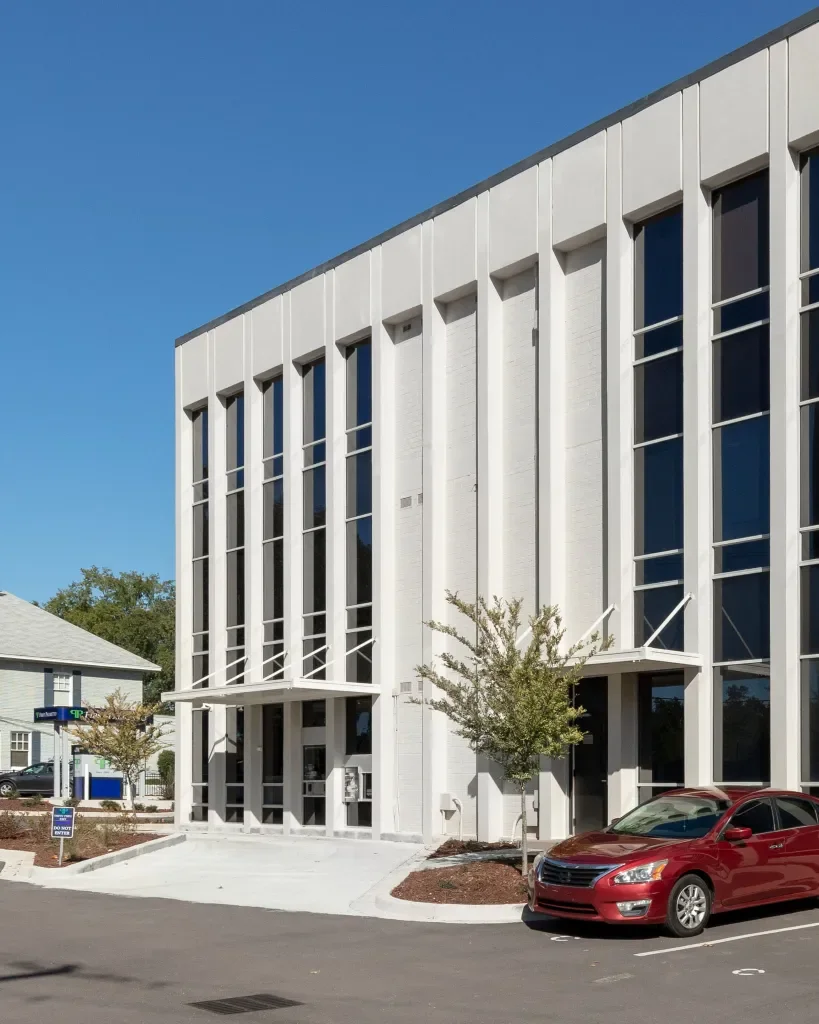 Modern white multi-story building with large vertical windows and small trees in front, parked red car, and a clear blue sky.