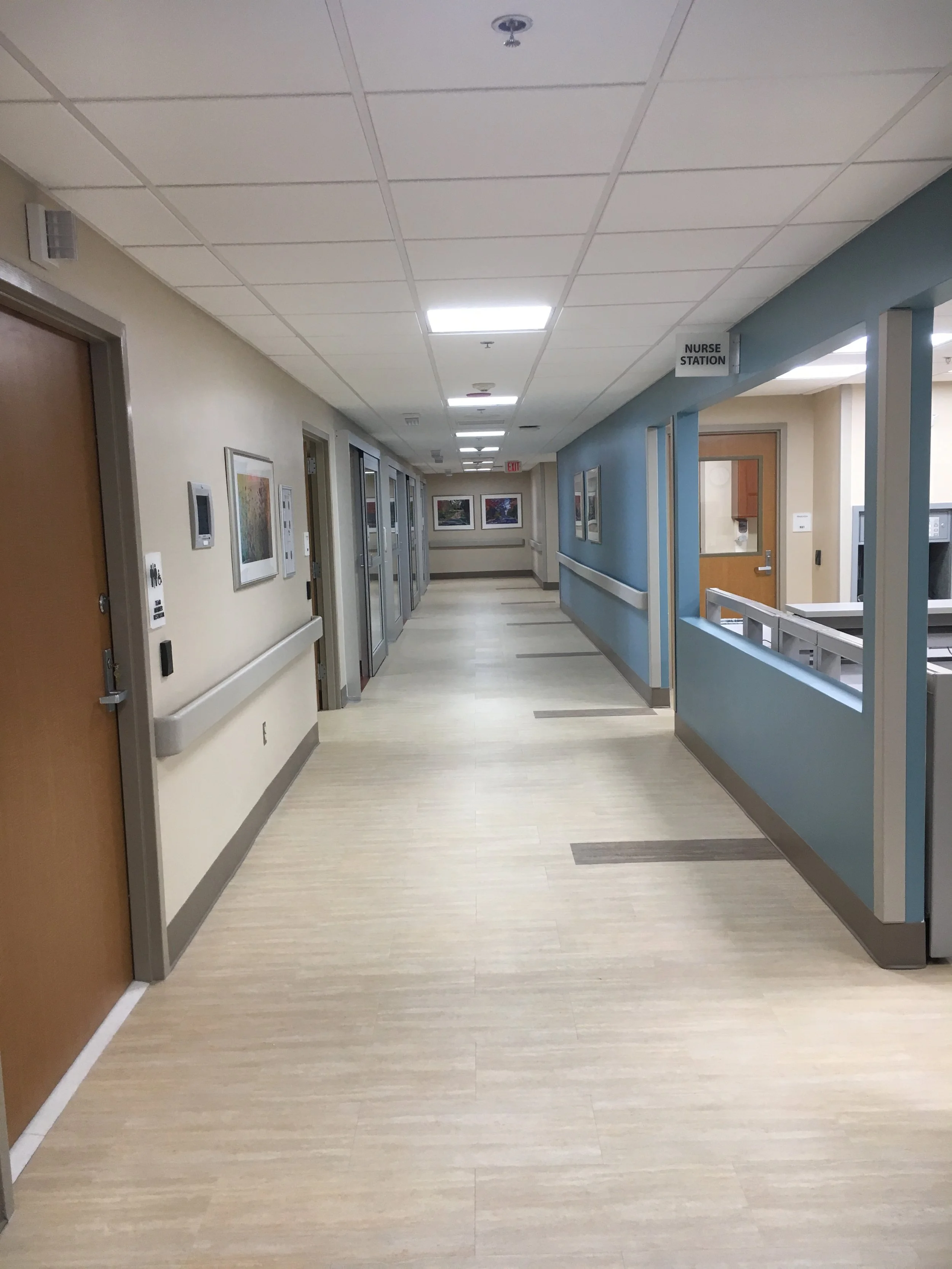 Hospital hallway with beige walls, framed artwork, elevator doors, and a nurse station visible through a blue wall opening.
