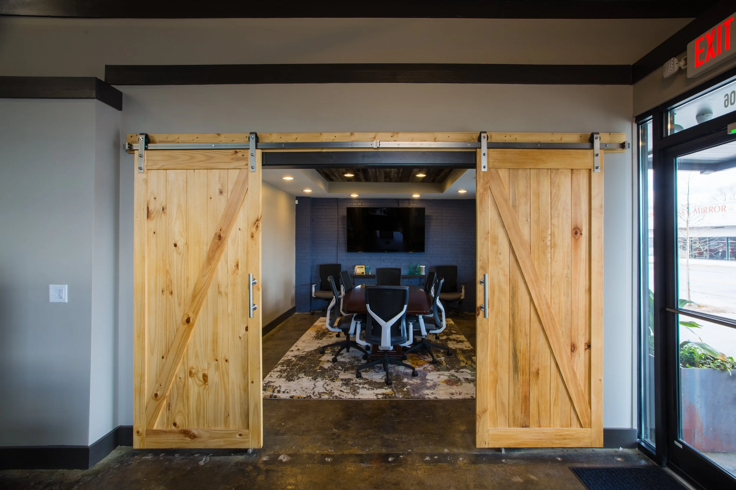 View through a sliding barn door into a conference room with a long table, multiple chairs, a television on the wall, and a decorative rug on the floor.