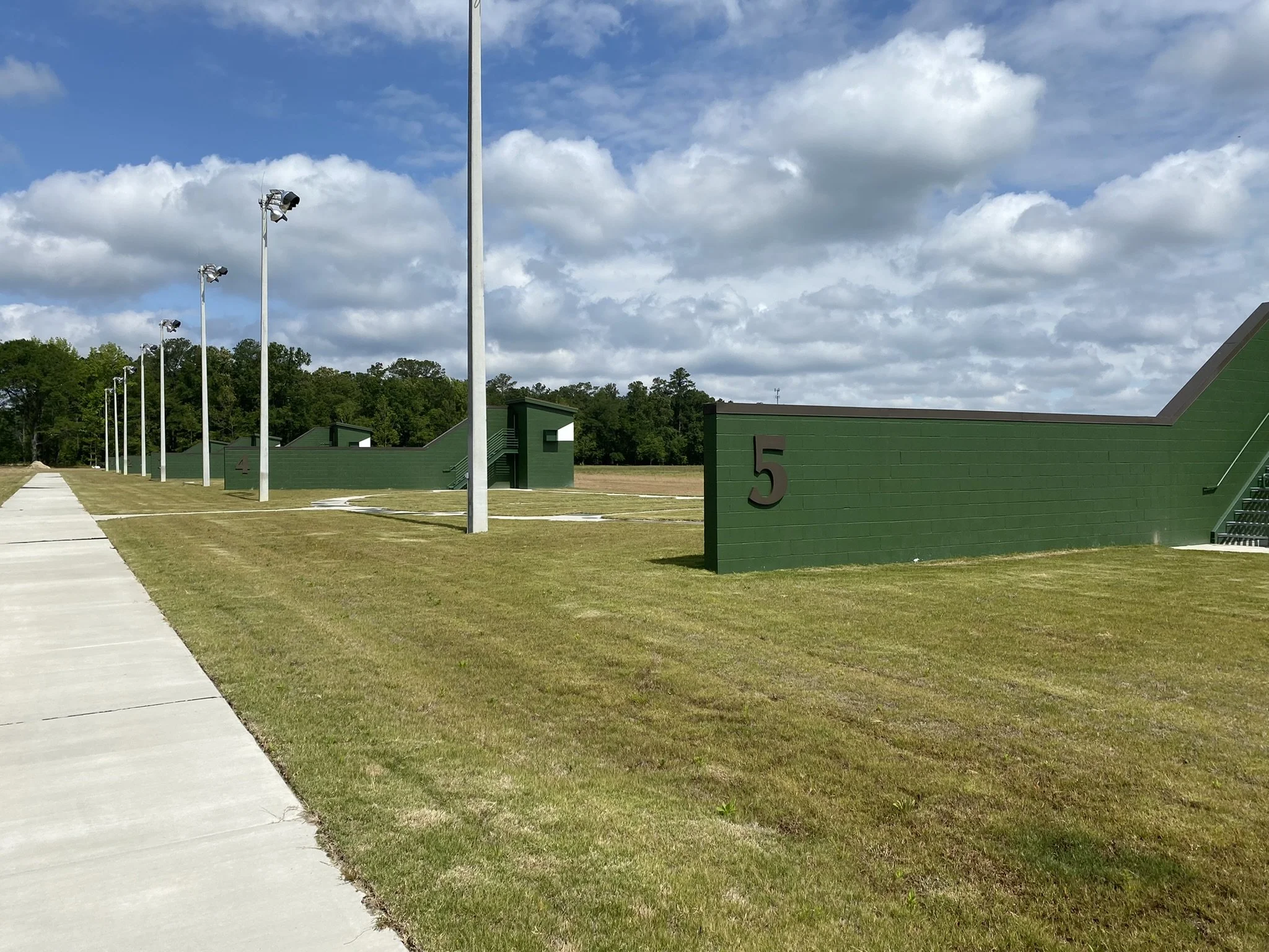 An outdoor sports facility with four green stadium-style structures labeled with numbers 4 and 5, tall light poles with multiple floodlights, a sidewalk, grass fields, and a partly cloudy sky.