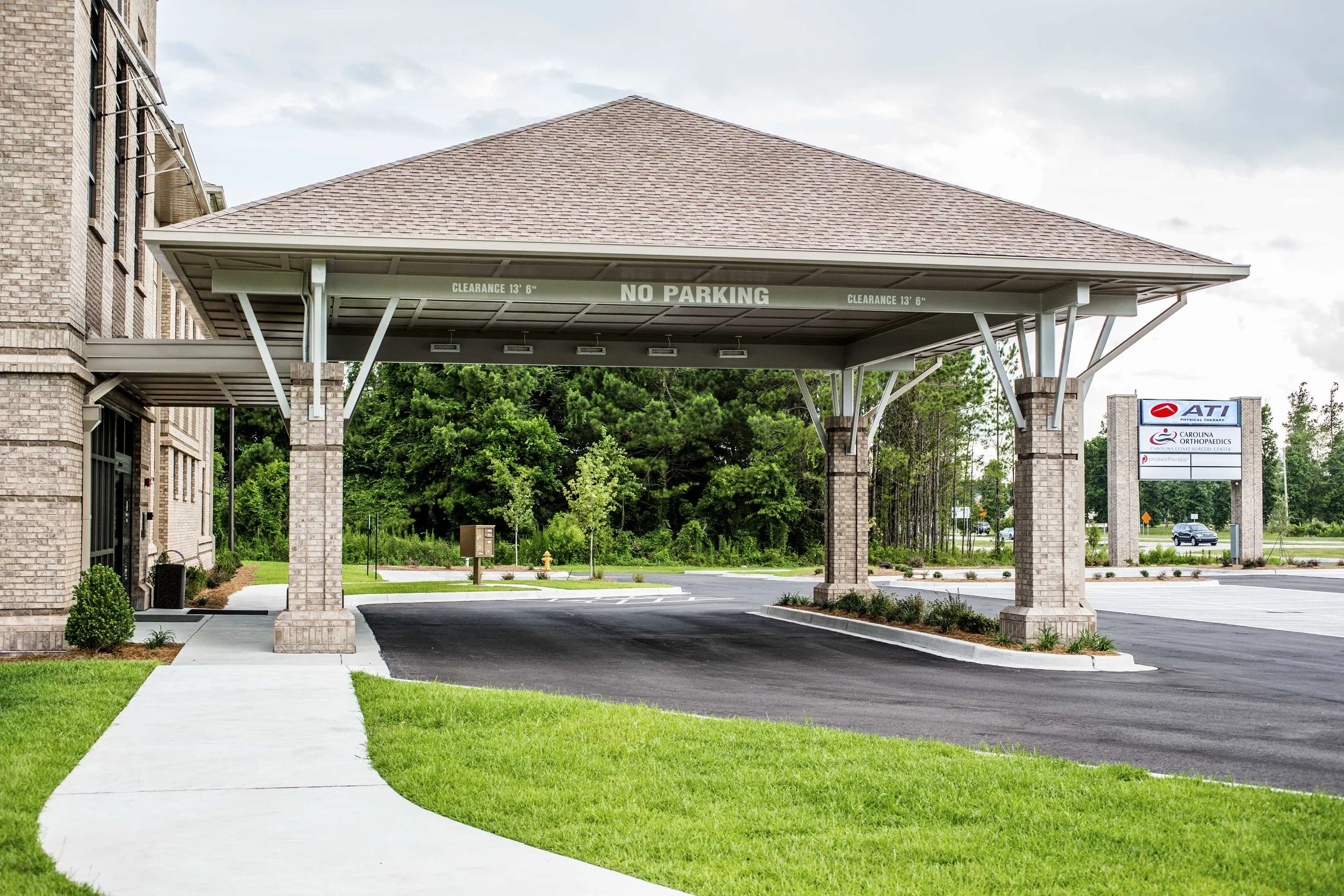Carport with brick pillars and a sign indicating 'No Parking' under a brown shingled roof, adjacent to a brick building and landscaped lawn.