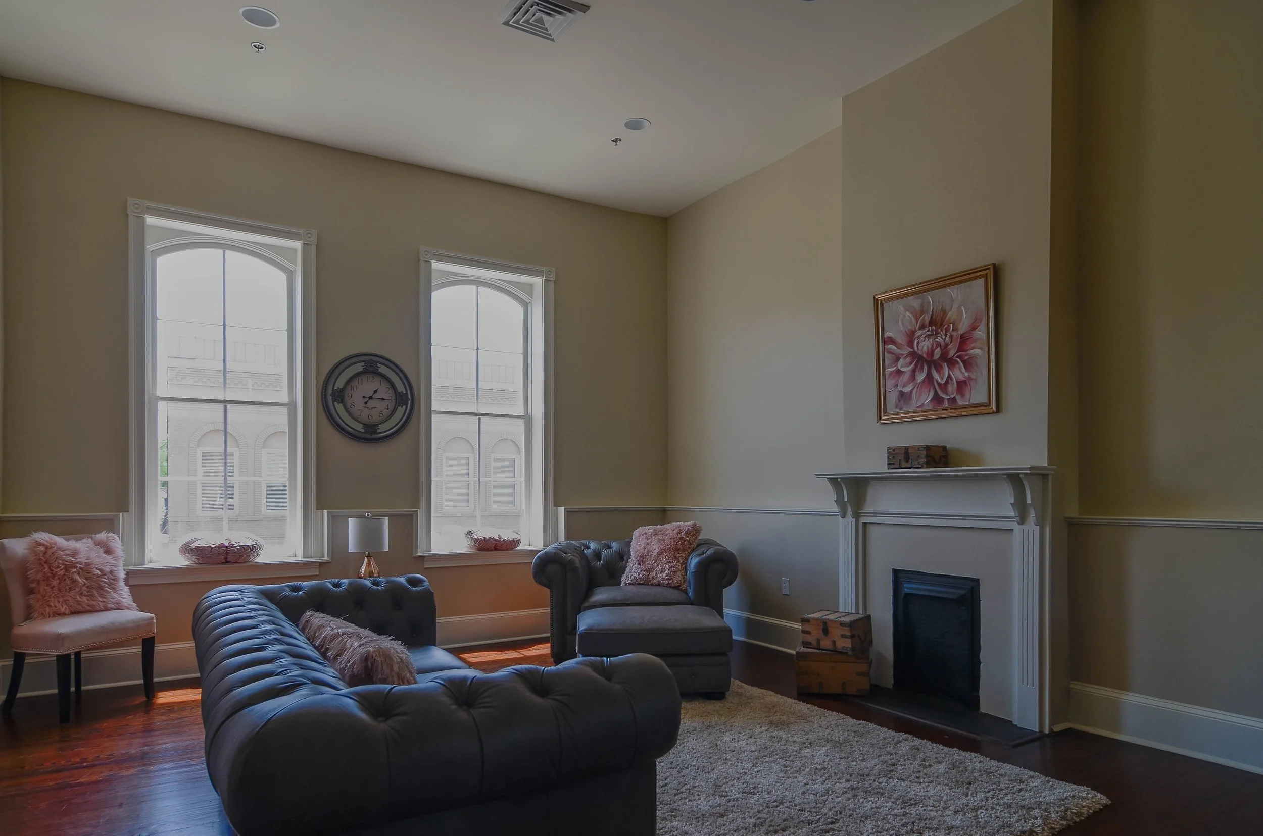 Living room with two tall windows, a white fireplace, a framed painting of a pink flower, a black leather tufted sofa, a gray armchair, pink decor pillows, a small side table with a white lamp, and a shaggy beige rug.
