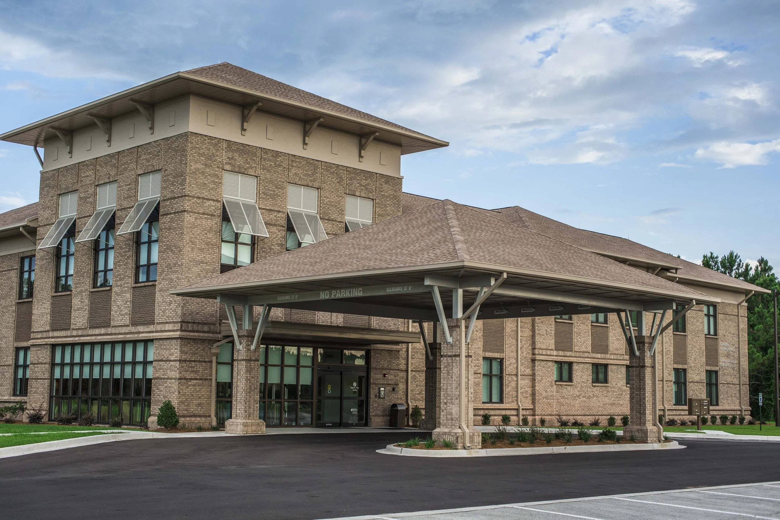 A brick multi-story building with large windows, a covered entrance, and a parking lot in front, under a partly cloudy sky.