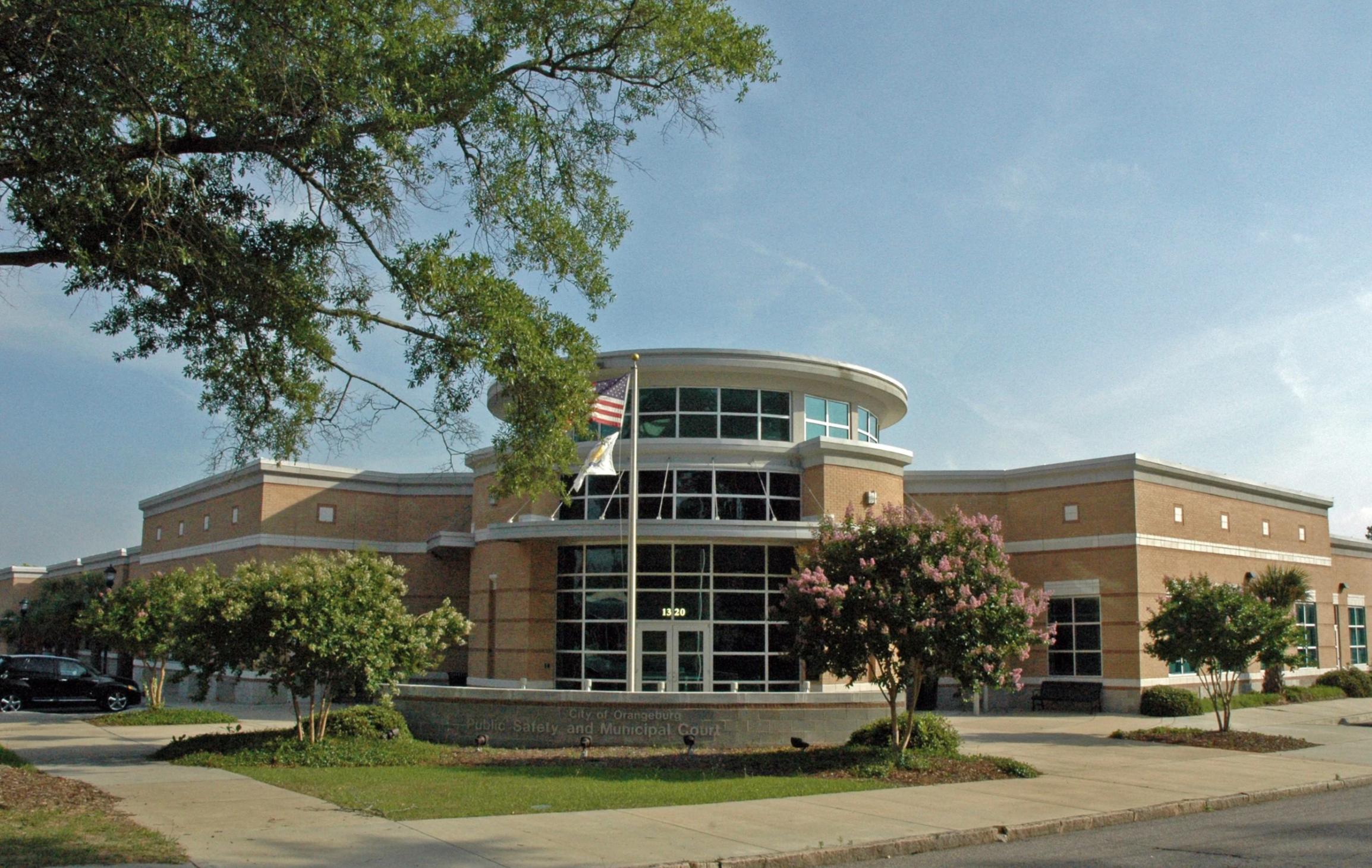 Front view of a county government building with a curved glass entrance, American flags, and well-maintained landscaping with trees and flowers under a blue sky.