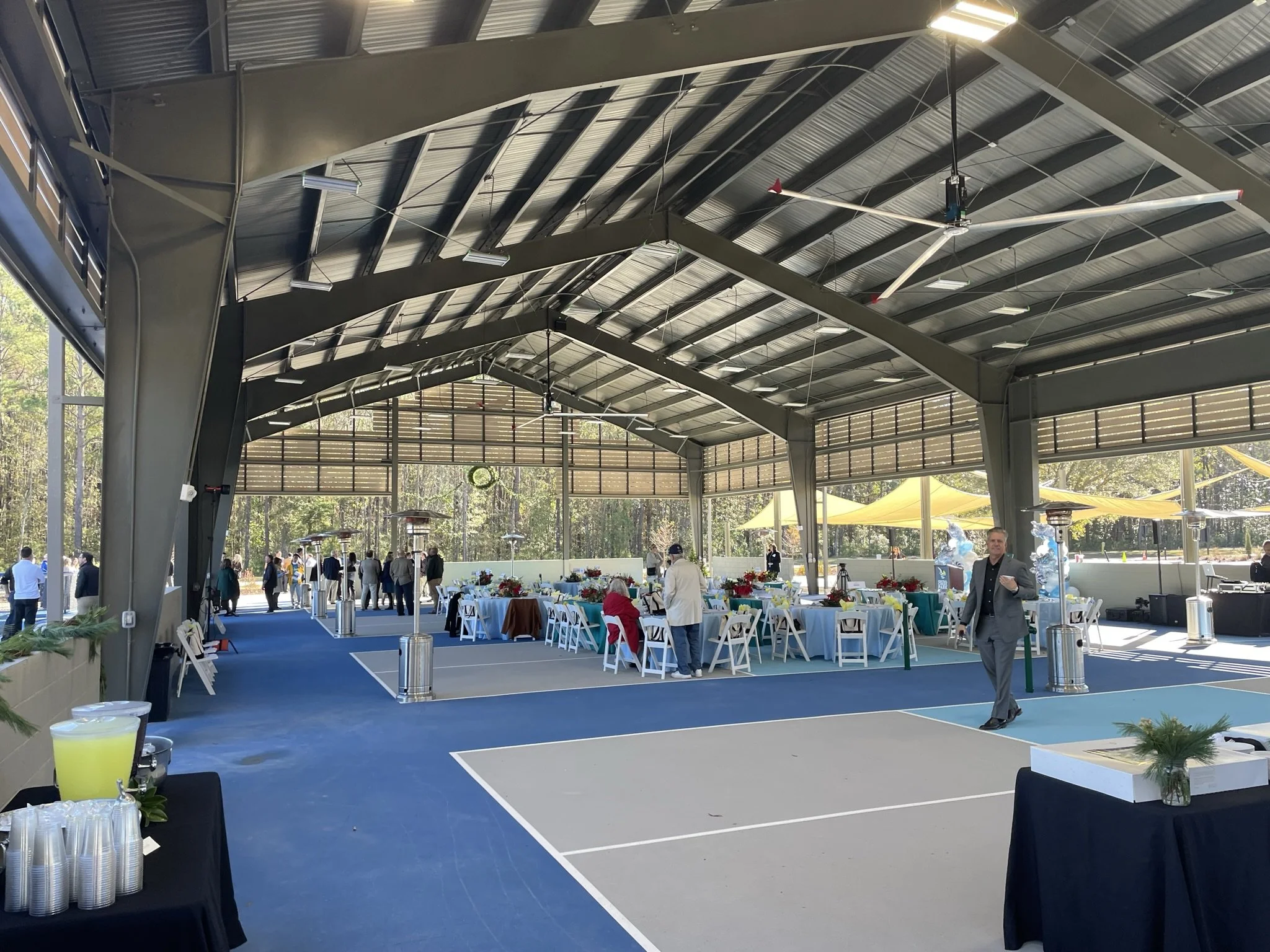An indoor event space with tables and chairs decorated for a celebration. The space has a high ceiling with metal beams and a ceiling fan. Outside, there are trees and yellow umbrellas visible through open windows. People are mingling and walking aro