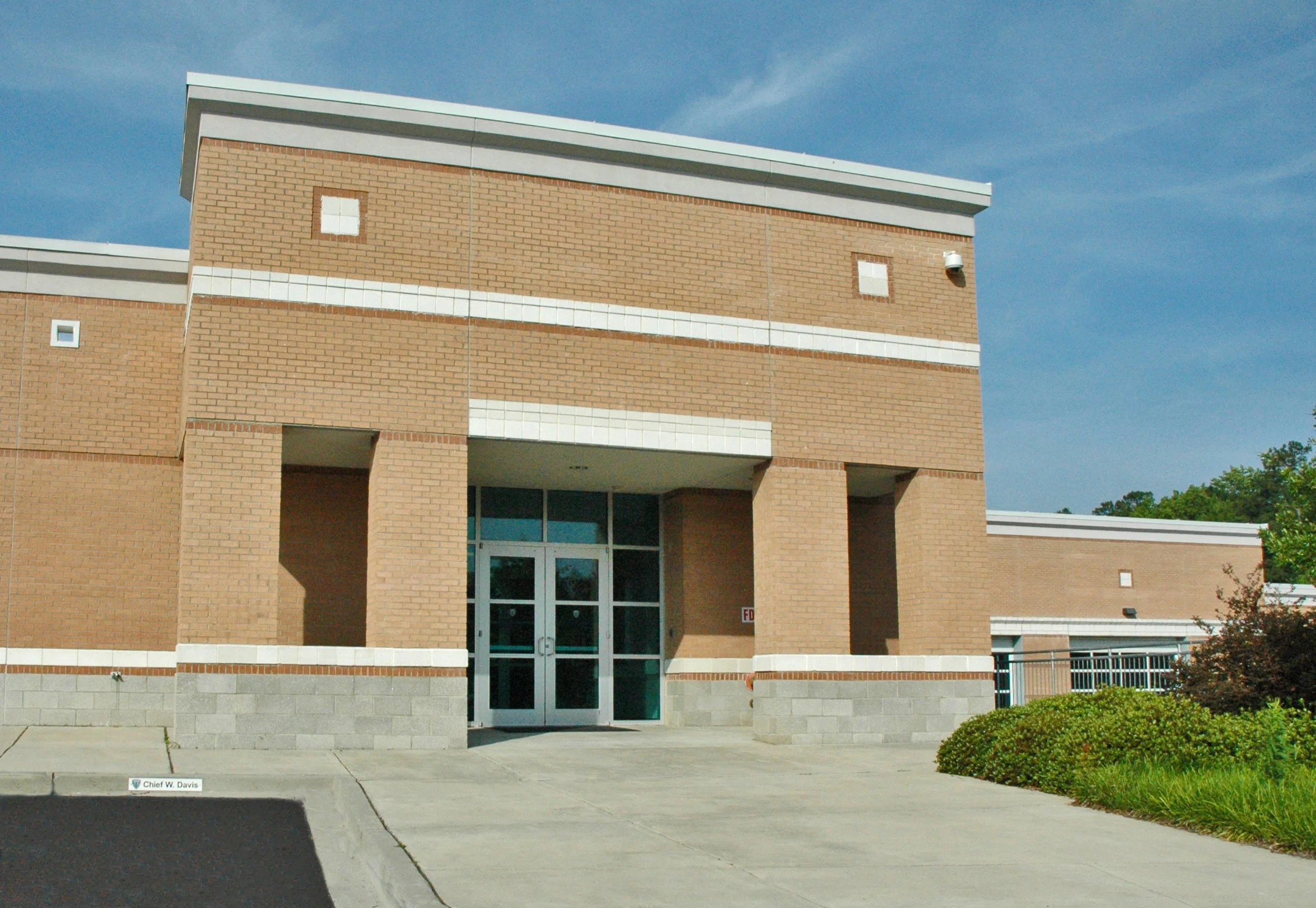 Facade of a brick school building with a glass entrance door, security camera, and surrounding bushes, under a partly cloudy sky.