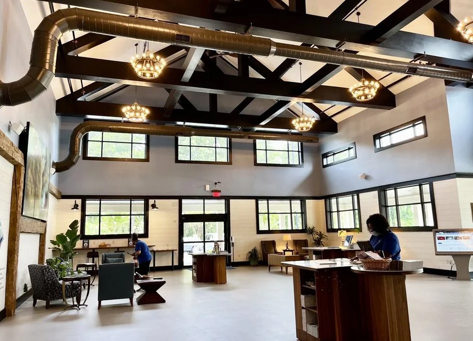 Modern, well-lit hotel lobby with high ceilings, large windows, and industrial-style lighting fixtures. Staff members are present near the reception desk.