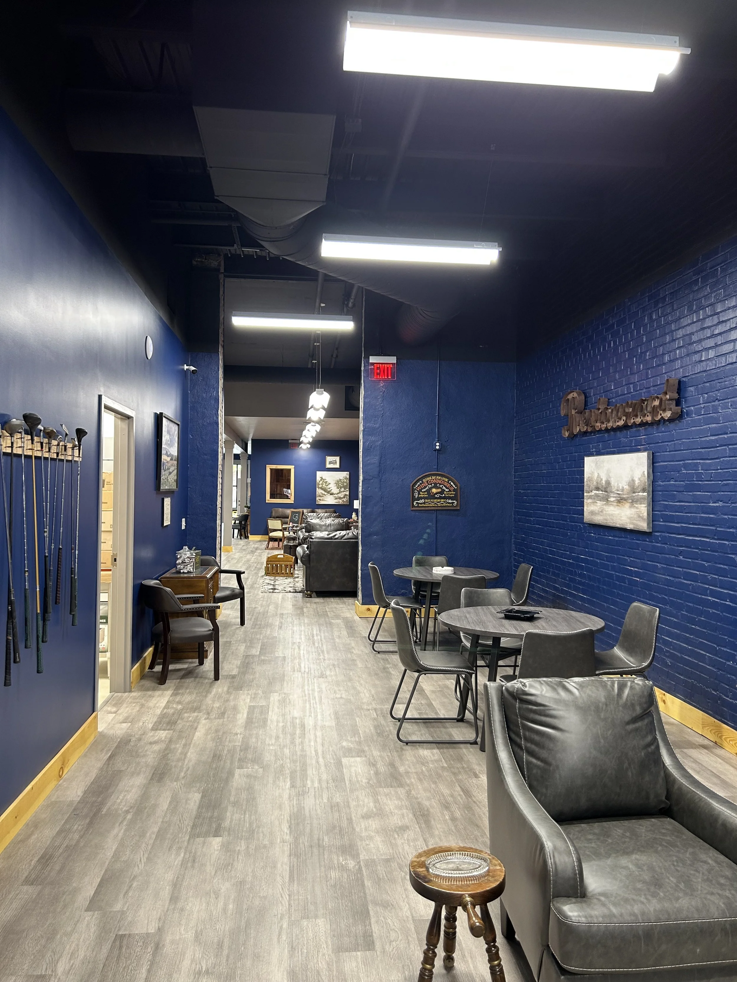 The image shows a hallway with blue walls and gray flooring, with furniture such as chairs and tables, paintings on the walls, and golf clubs mounted on the wall to the left. There is an exit sign and various lights on the ceiling.