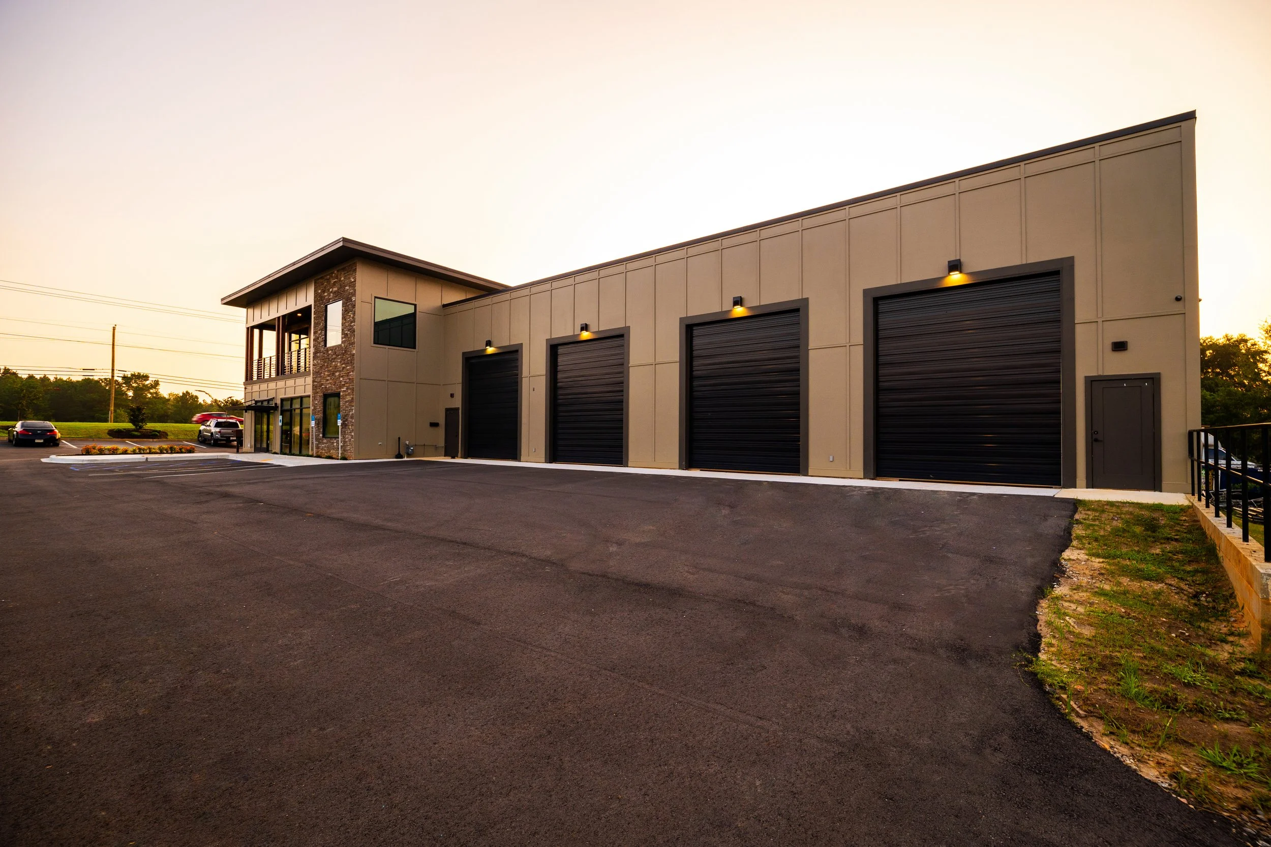 A modern commercial building with three large black garage doors illuminated by exterior lights, a small parking lot with a few cars, and a landscaped area with flowers, all under a clear sky at sunset.