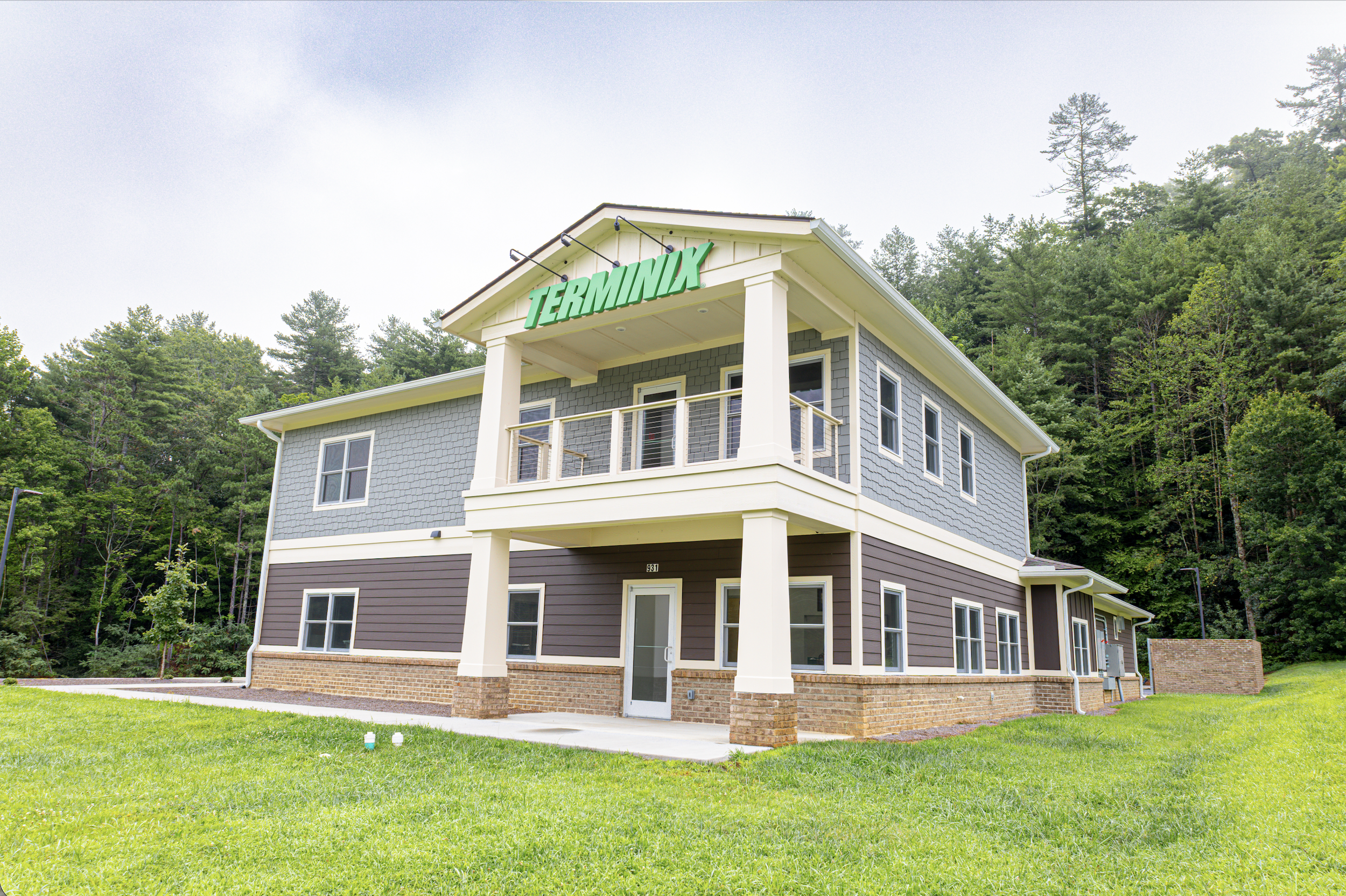 A two-story house with grey siding and white trim, featuring a green sign with the word 'TERMINIX' on the upper front of the house, surrounded by green grass and trees in the background.