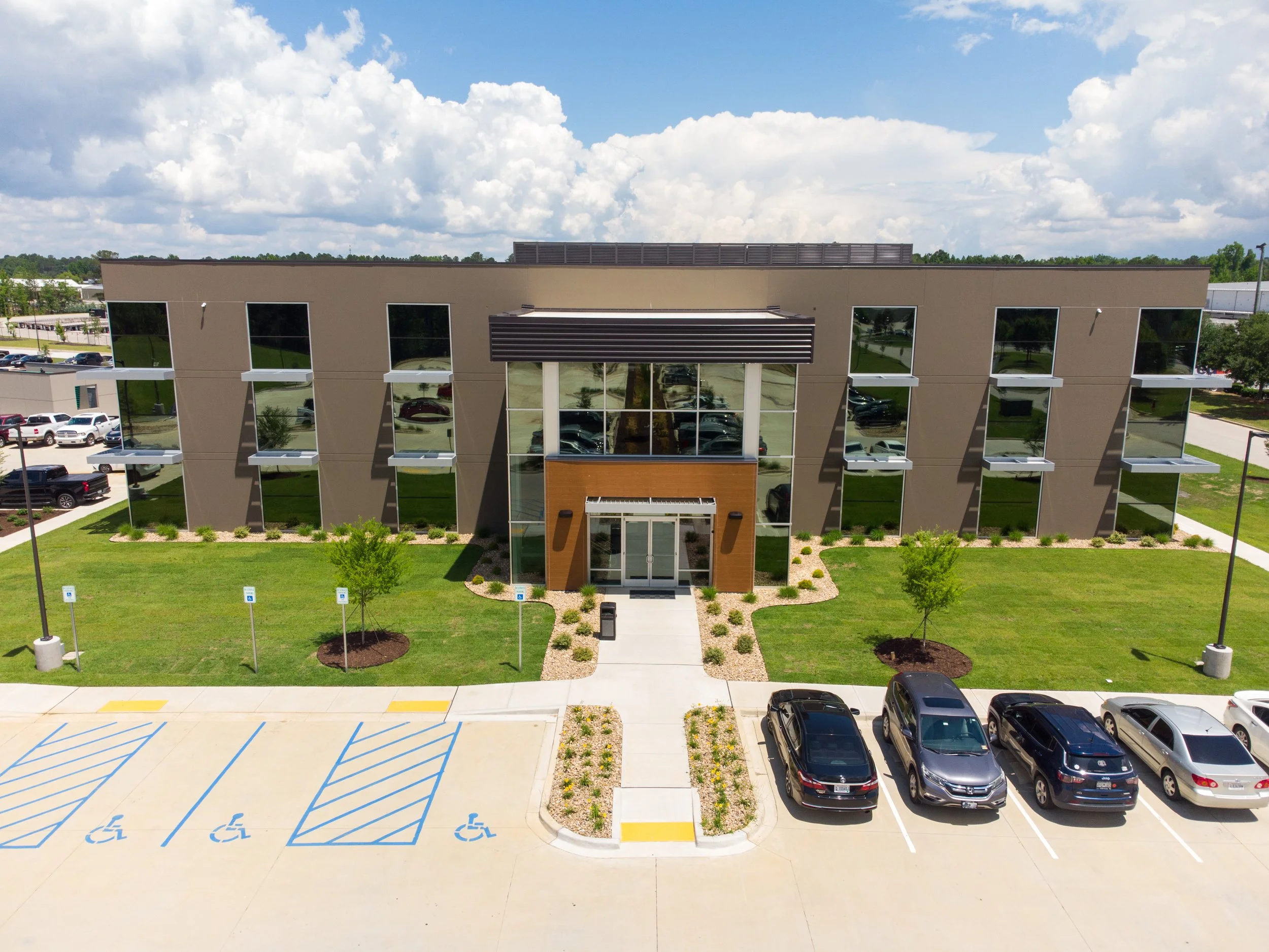 An office building with reflective glass windows and a modern design, surrounded by a parking lot with several cars, green grass, and small trees, under a partly cloudy sky.