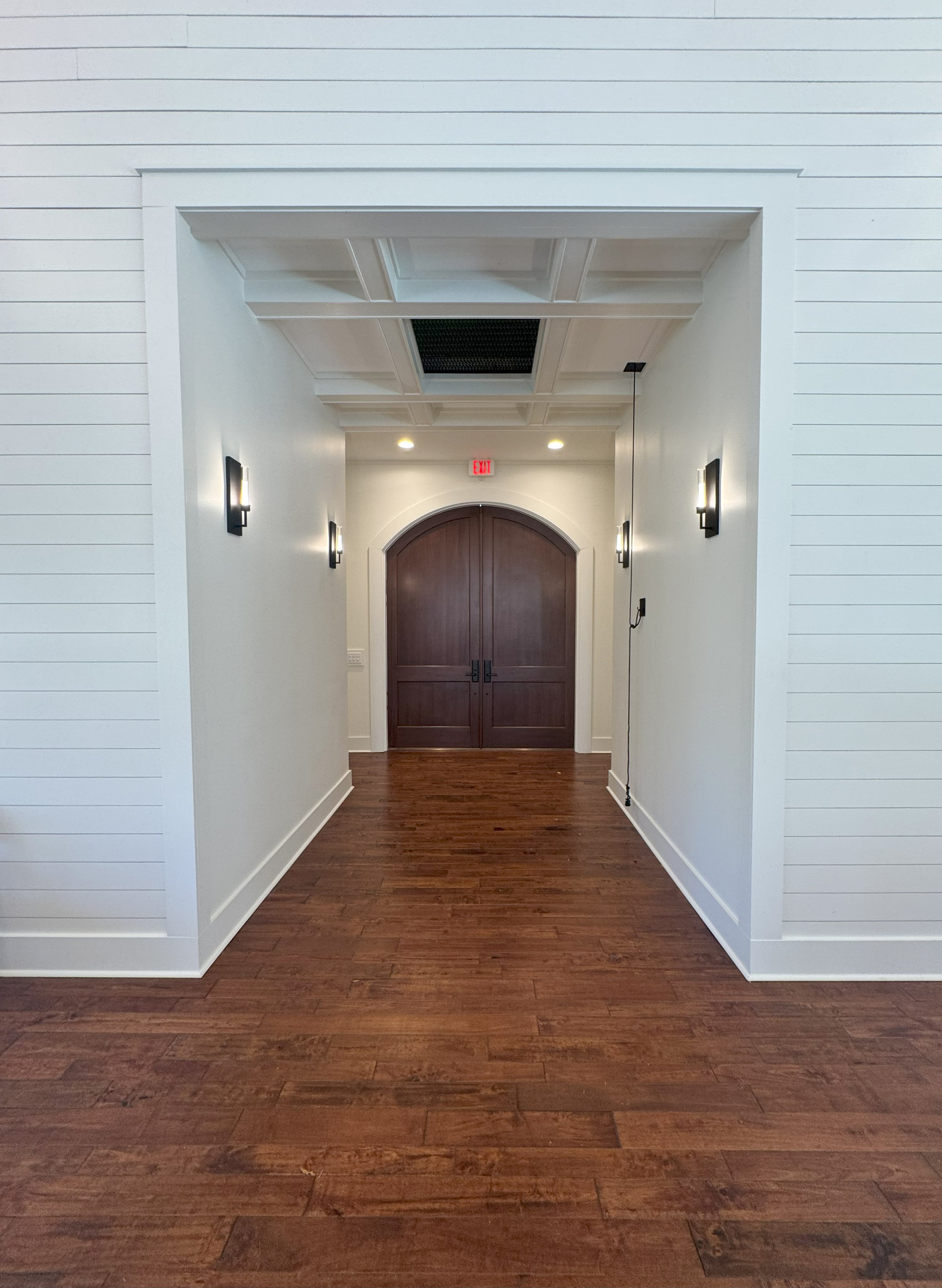Interior hallway with white walls, dark wood double doors, and wooden flooring, lit by wall sconces and ceiling lights.