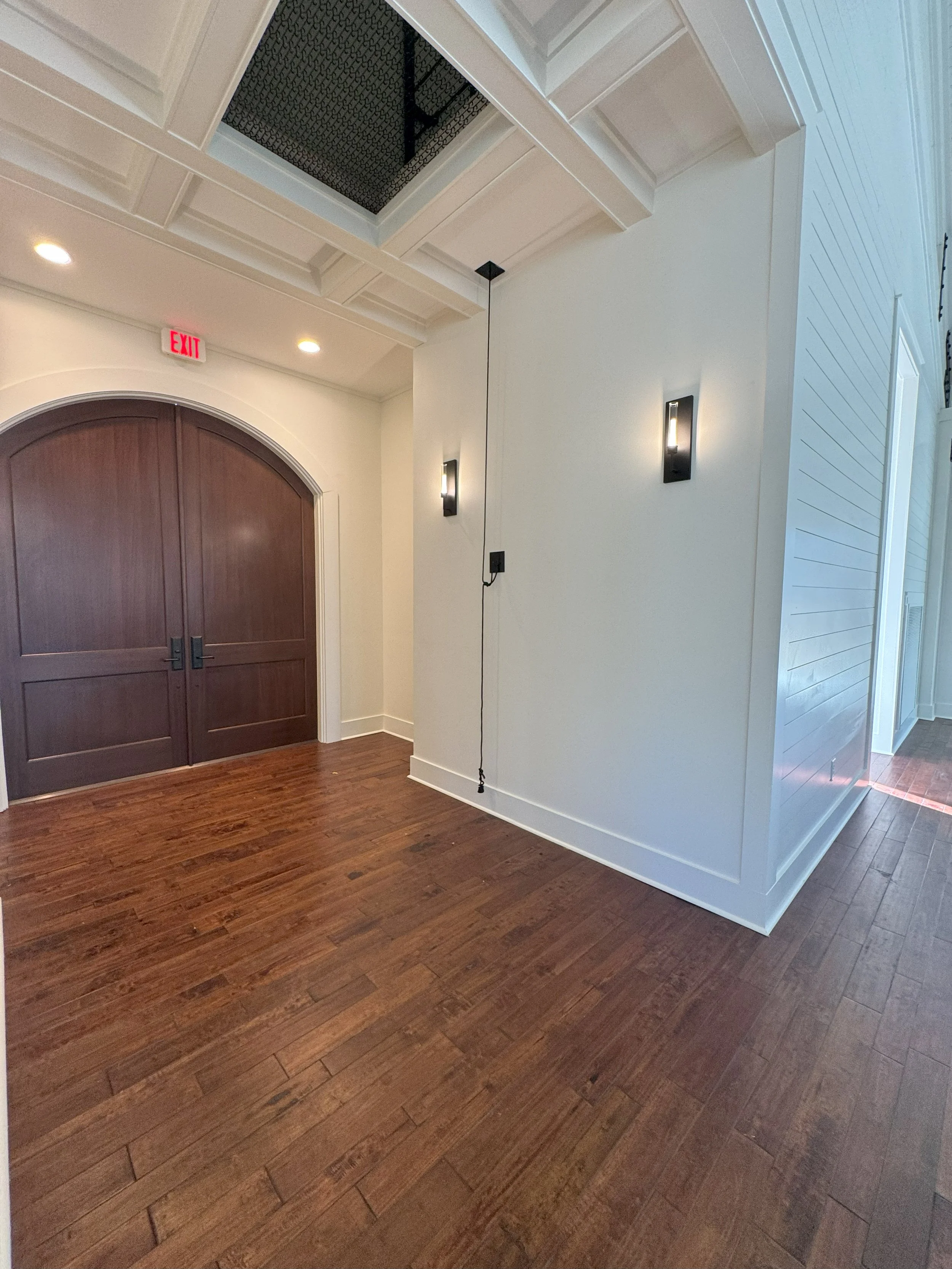 Interior view of a room with a wooden floor, a white wall with two black wall-mounted lights, a ceiling with decorative molding, a ceiling vent with a black cover, and a large arched wooden double door.