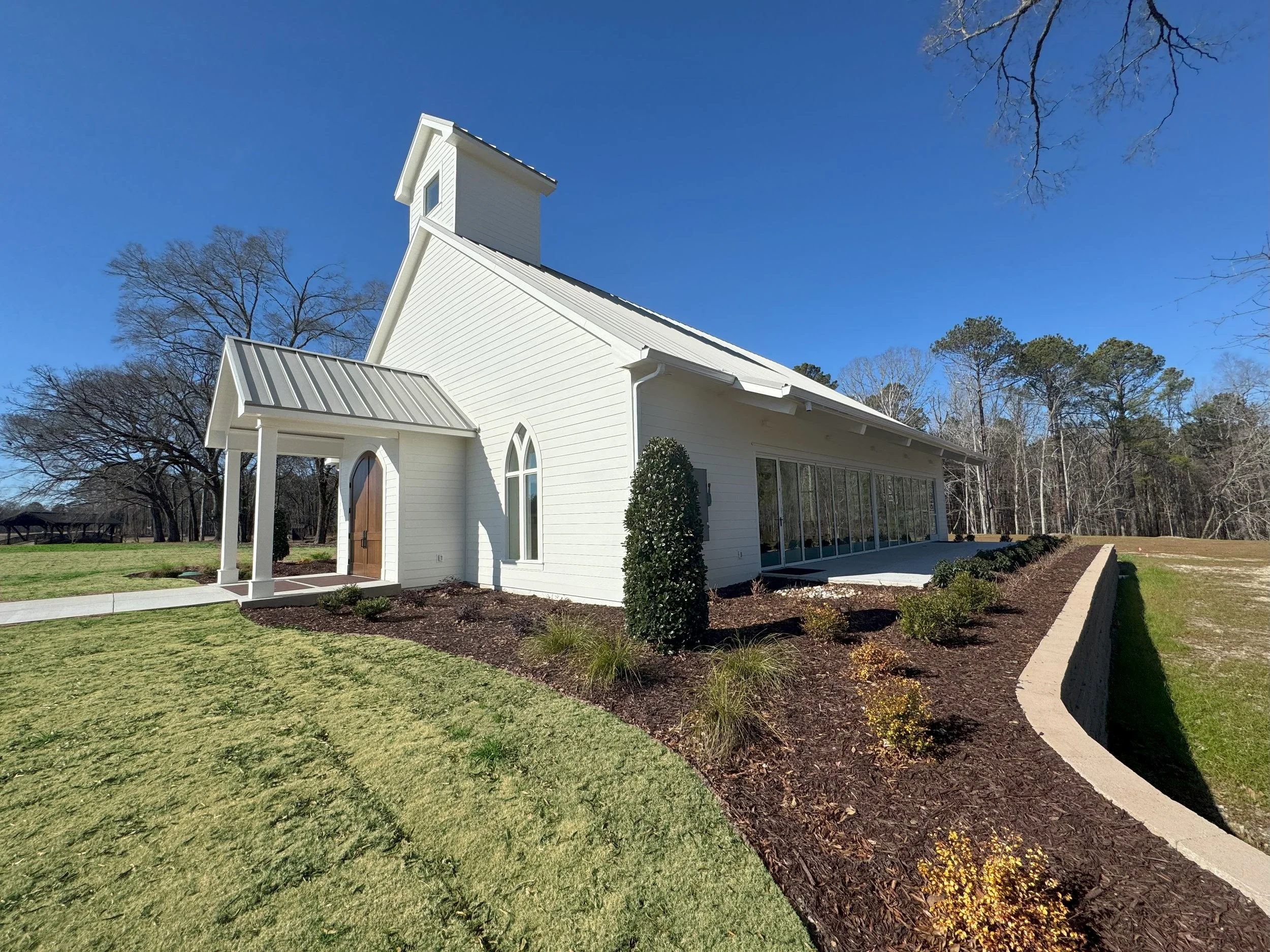 A white church with a metal roof and steeple, surrounded by trees and a well-maintained lawn under a clear blue sky.