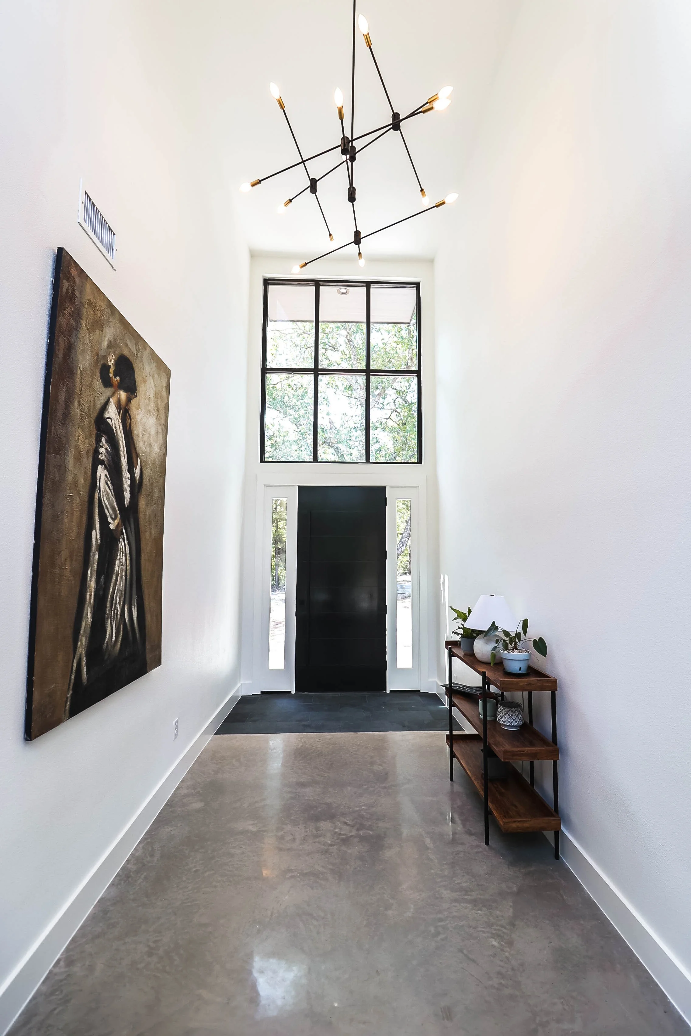 A modern entryway with a black front door, large window above, white walls, a concrete floor, and a wooden and metal console table with plants and decorative objects on the right. A large artwork of a woman hangs on the left wall, and a black contemp