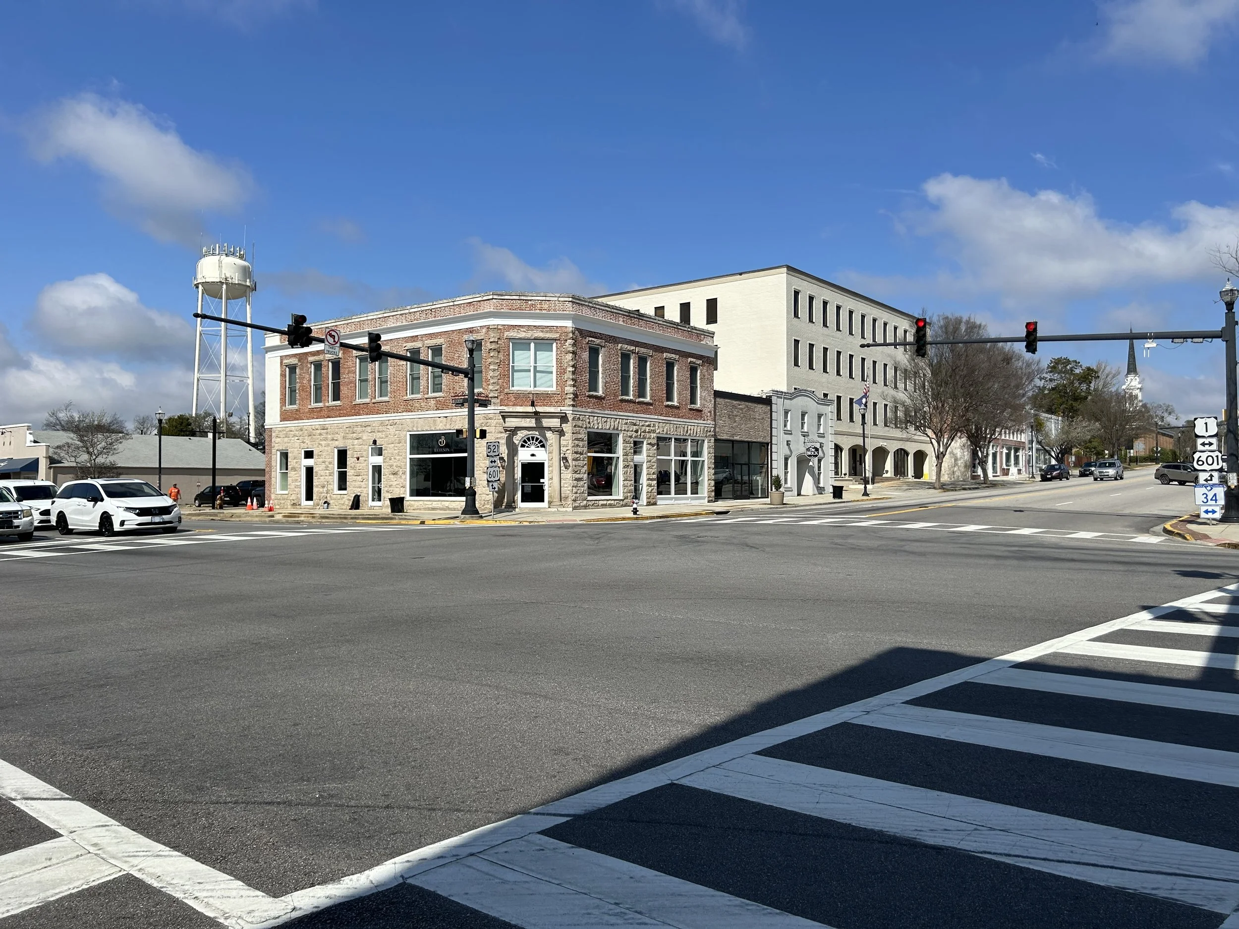 Downtown street scene with cars parked along the curb, traffic lights, historic brick and stone buildings, and a partly cloudy blue sky.