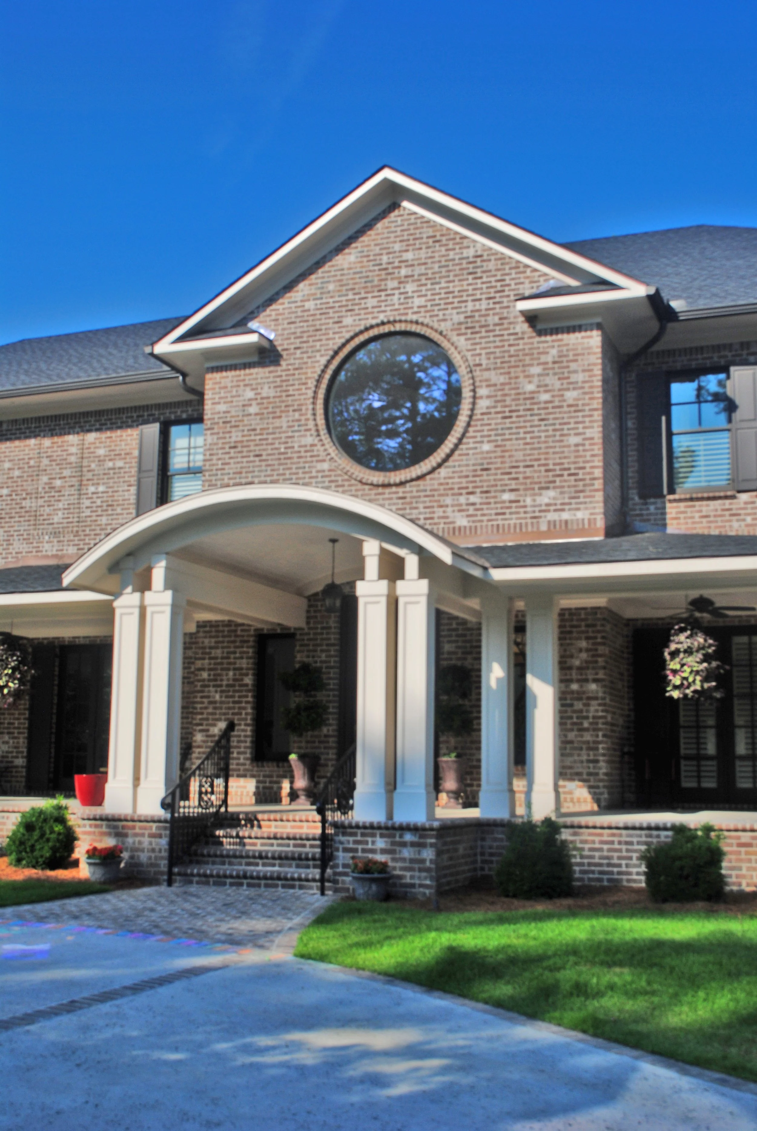 Front view of a brick house with a circular window and a covered porch supported by white columns, with a well-maintained lawn and a driveway.
