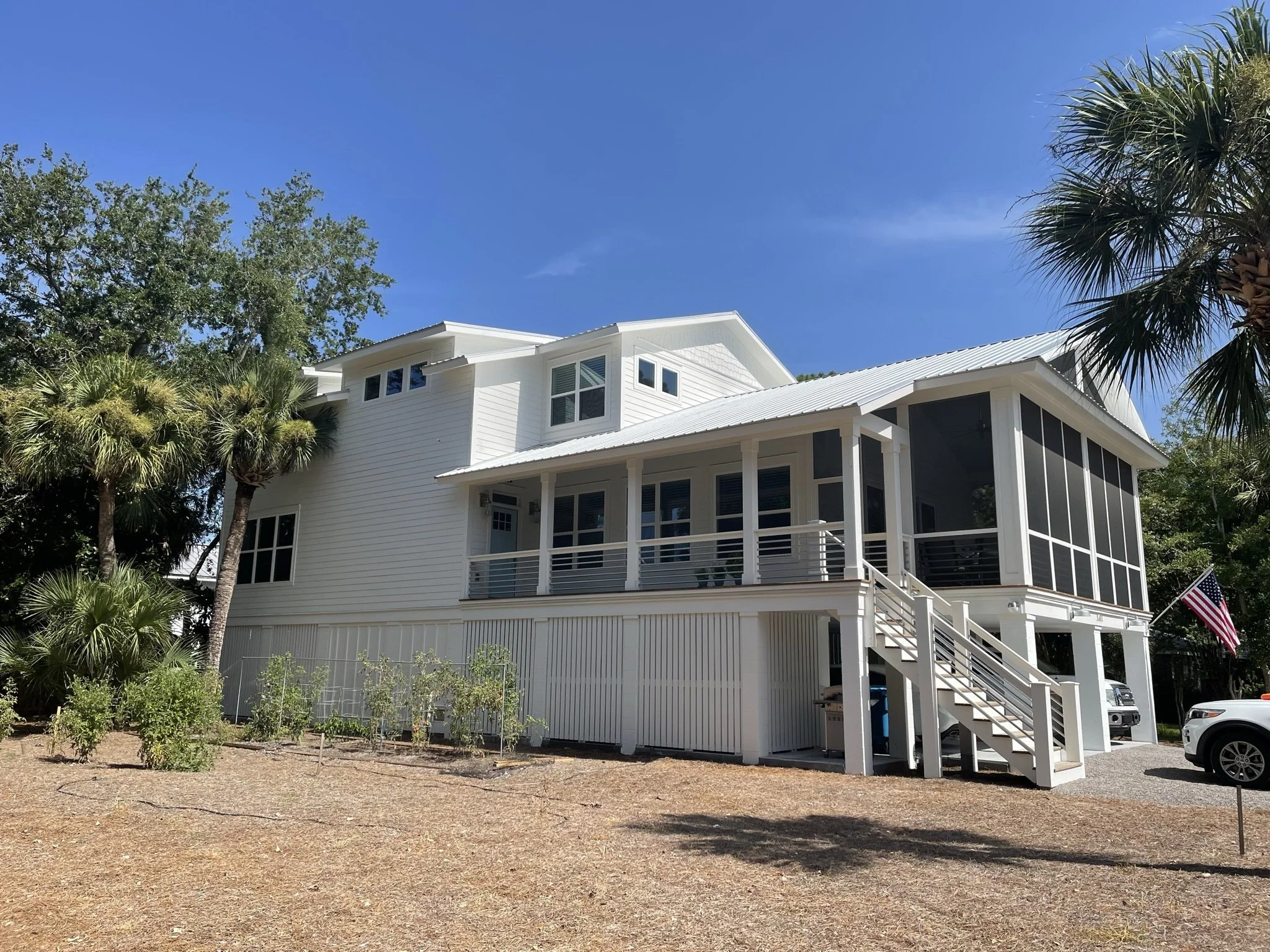 A white house with a metal roof, surrounded by trees and plants, with a porch and a staircase leading to the entrance. The sky is clear and blue.