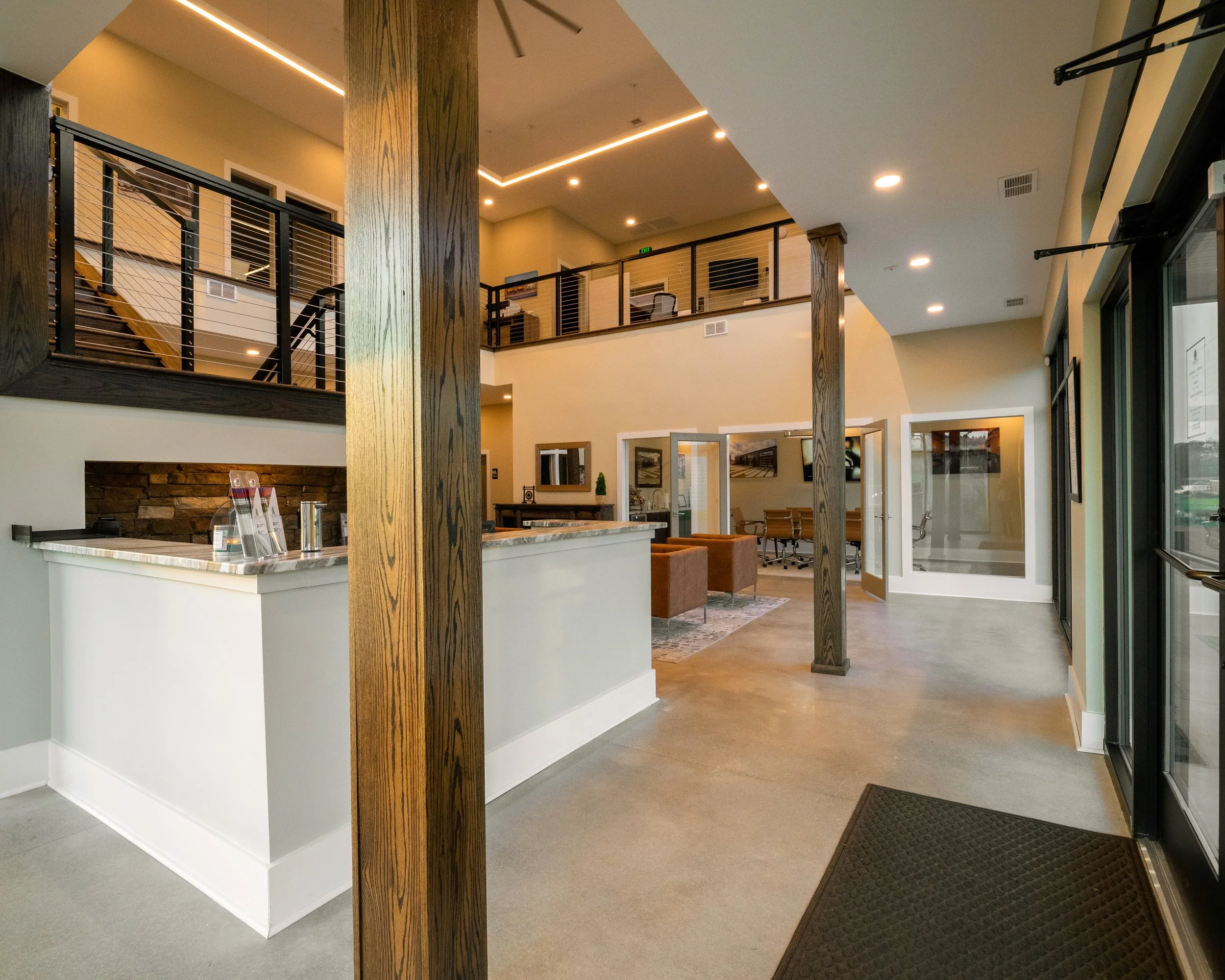 Interior view of a modern office or commercial space with open layout, featuring wooden columns, a reception counter, seating area, and a second-floor balcony with office desks and chairs overlooking the main area, illuminated by ceiling lights.
