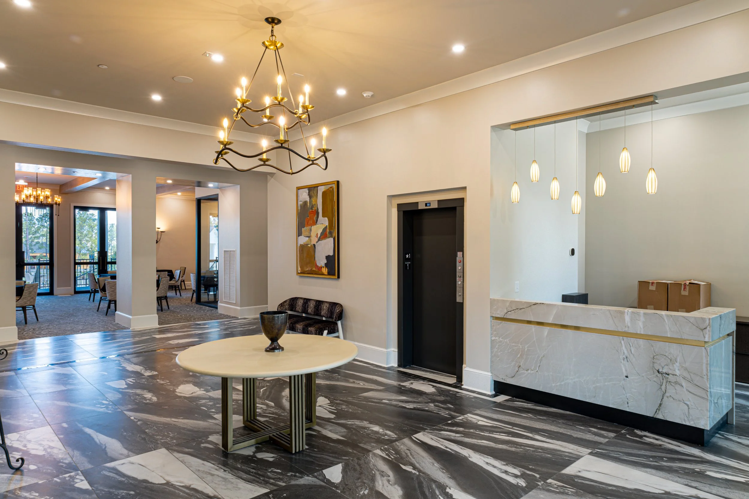 Interior view of a modern hotel lobby with a circular marble table with a decorative black bowl, a patterned black and white floor, a gold chandelier hanging from the ceiling, and a reception desk with a white marble front and gold accents. There are