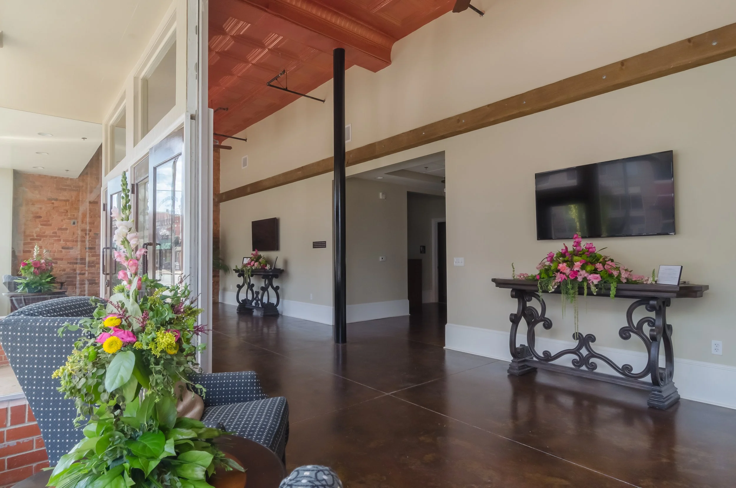 Interior of a room with flowers on tables, a television on the wall, brick and beige walls, wooden beams, and a wooden floor.