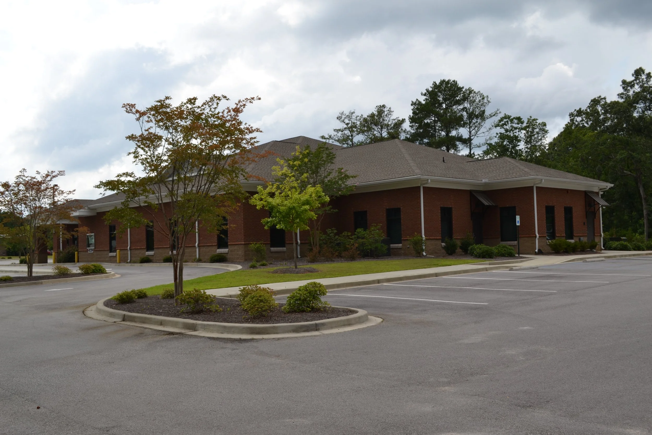 Empty parking lot with a few trees and a brick building in the background under a cloudy sky.