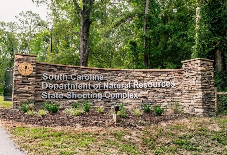 Stone entrance sign for South Carolina Department of Natural Resources State Shooting Complex, surrounded by landscaped plants and trees.