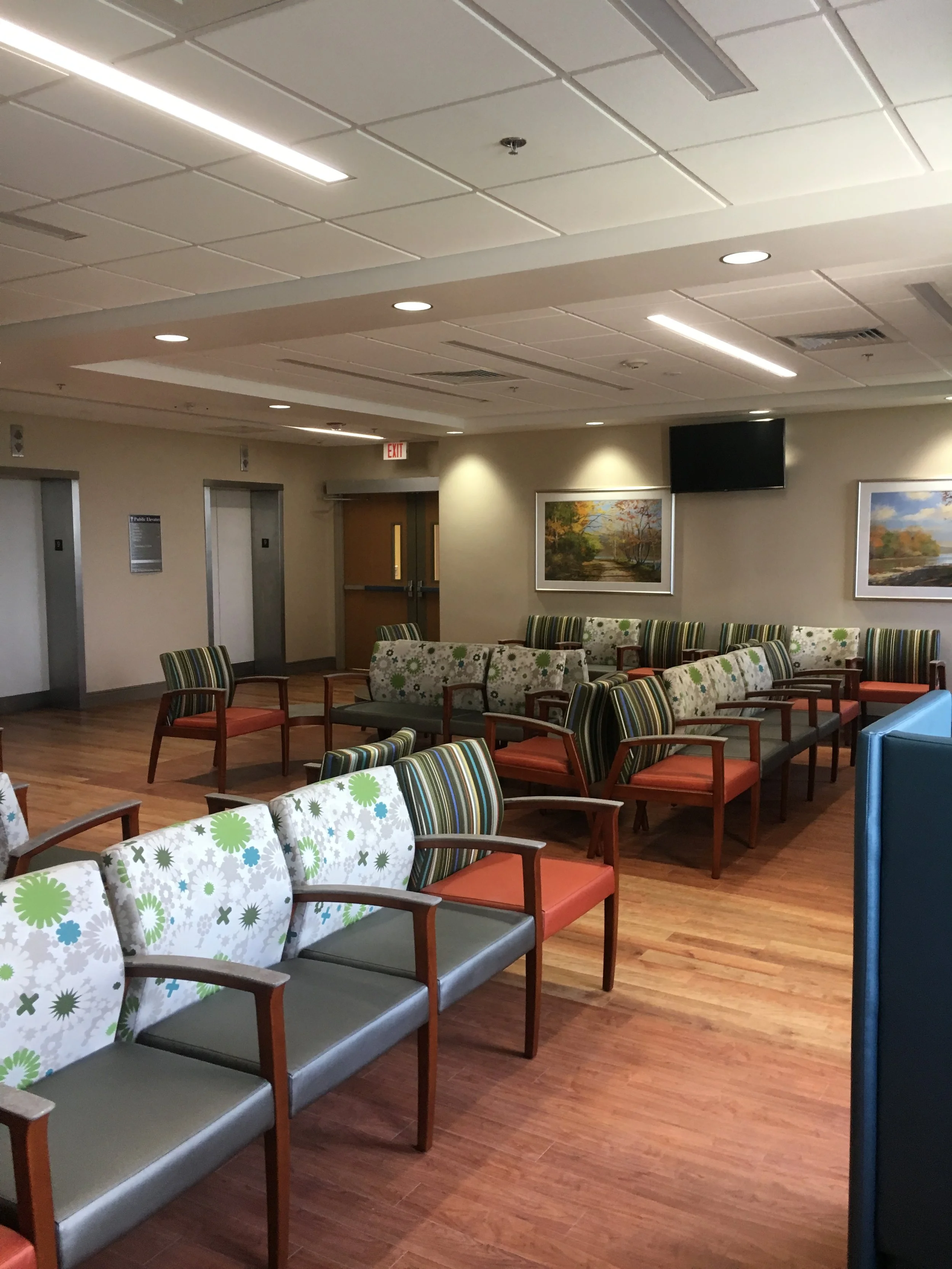 A waiting area with several chairs arranged in rows, some with colorful striped or floral cushions, in a room with a wooden floor, paintings on the wall, a television, and elevator doors.