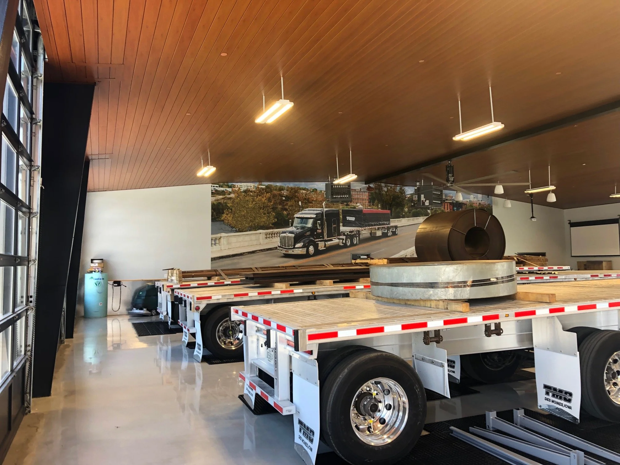 A truck loader inside a building with a wood-paneled ceiling, bright lighting, and large windows showing an outdoor parking lot with trucks and trees.