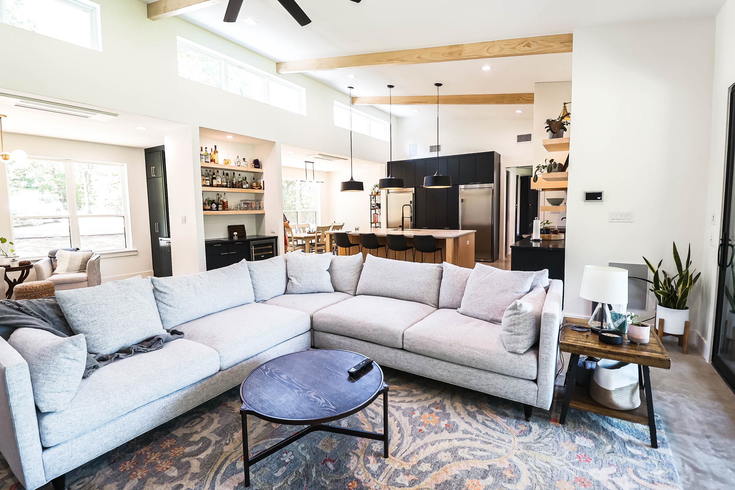 A modern open-plan living room and kitchen with large windows, a light-colored sectional sofa, a black coffee table, and a patterned area rug. The kitchen features black cabinetry, a large island with bar stools, hanging pendant lights, and built-in 