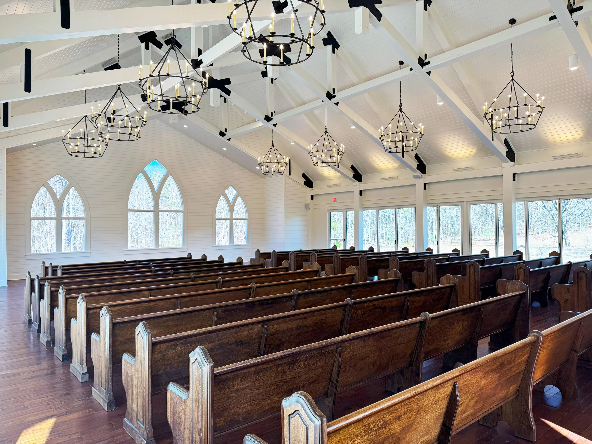 Interior of a church with wooden pews, large windows, and chandeliers hanging from the ceiling.