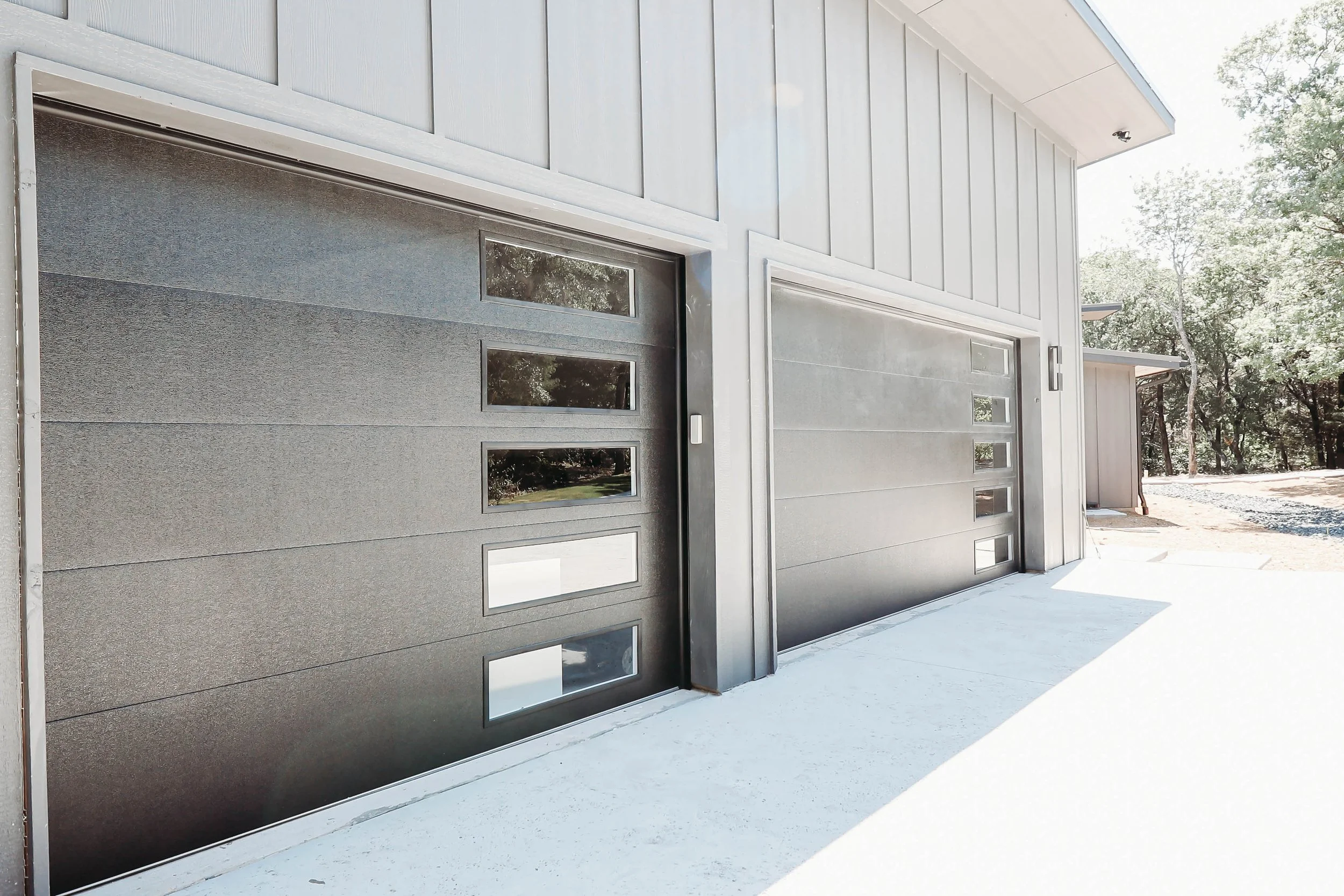 Two modern garage doors with small rectangular windows, part of a light-colored house exterior, with some trees in the background.