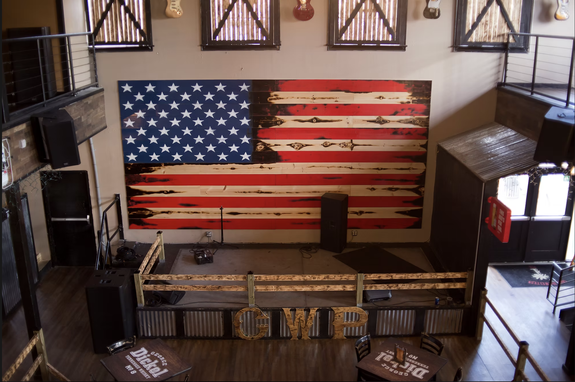 Interior of a bar or restaurant decorated with a large American flag made of wood on the wall. There are guitars hanging above the flag, a TV on the left, and a railing on the upper level. The space has tables with beer branding and a small stage are