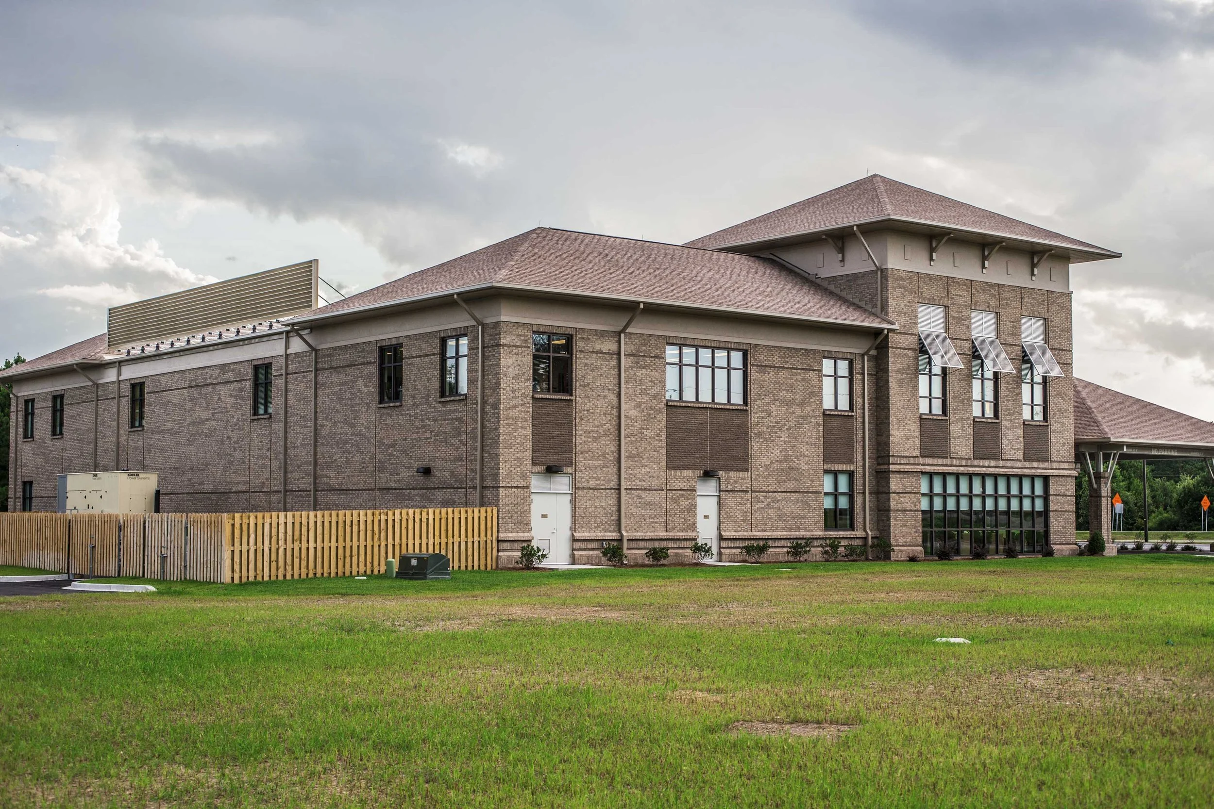 A three-story brick building with large windows, a pitched roof, and a fenced yard, under a cloudy sky.