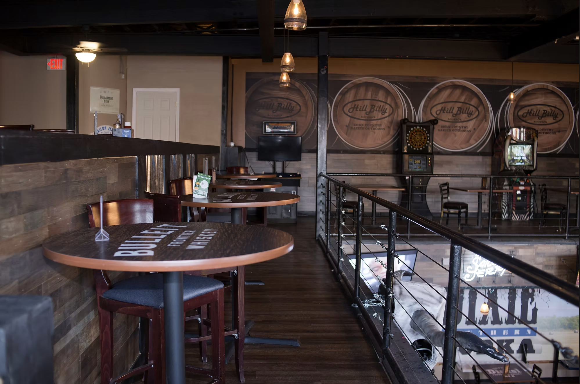 Interior of a restaurant or bar with wooden accents, circular tables, chairs, darts game, and a mural of wood barrels with the logo 'Hill Billy' on the wall.