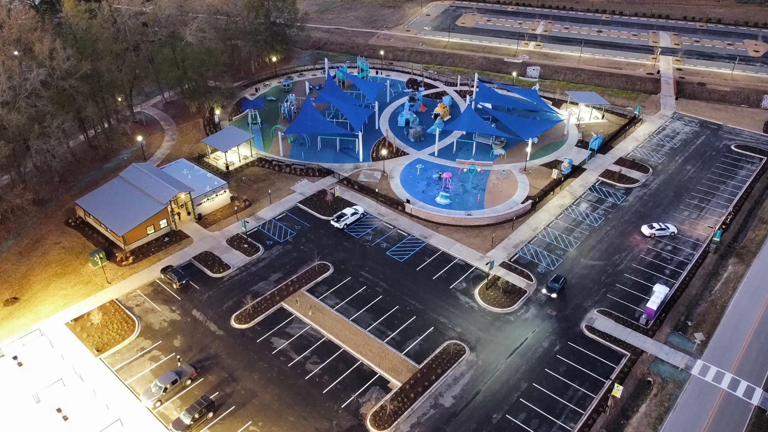 An aerial view of a brightly lit outdoor playground at night, with multiple blue shade canopies, playground equipment, and surrounding parking lots with several cars parked, nearby trees, pathways, and small structures.