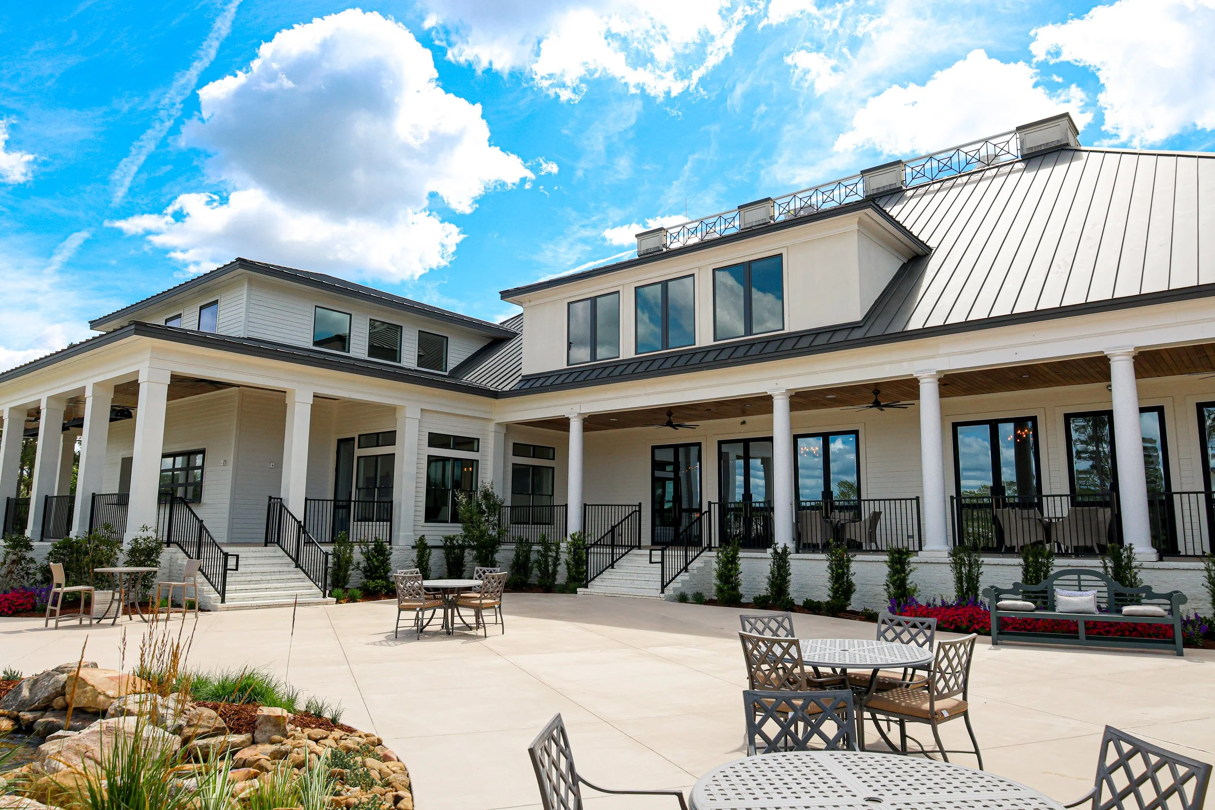 A modern white house with large windows and a metal roof, with a spacious outdoor patio area featuring metal tables and chairs, and landscaped garden beds, under a partly cloudy sky.