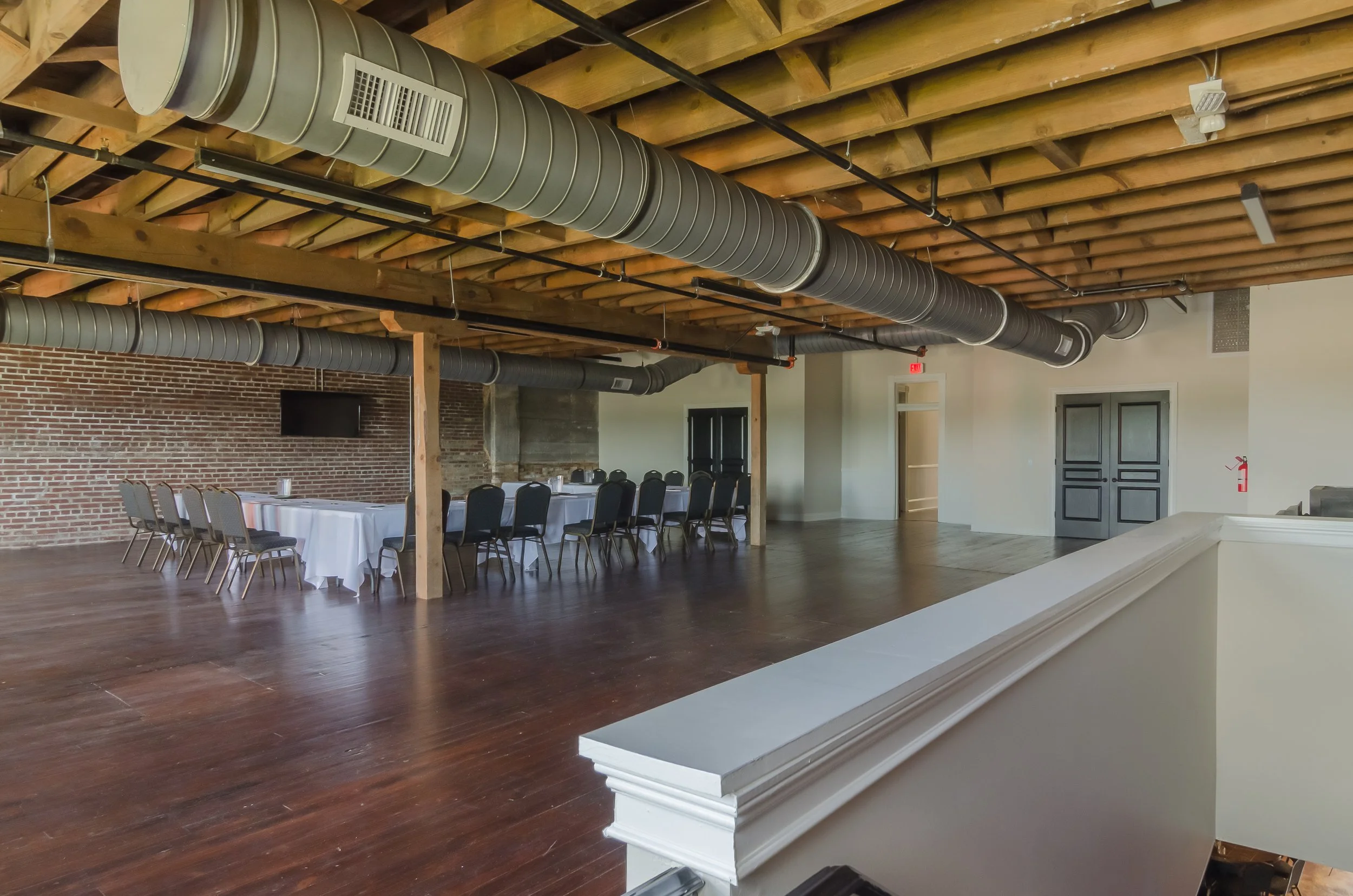 An unfinished indoor event space with exposed wooden ceiling beams and ductwork, brick wall, and a long table with chairs, indicating a rustic or industrial style.