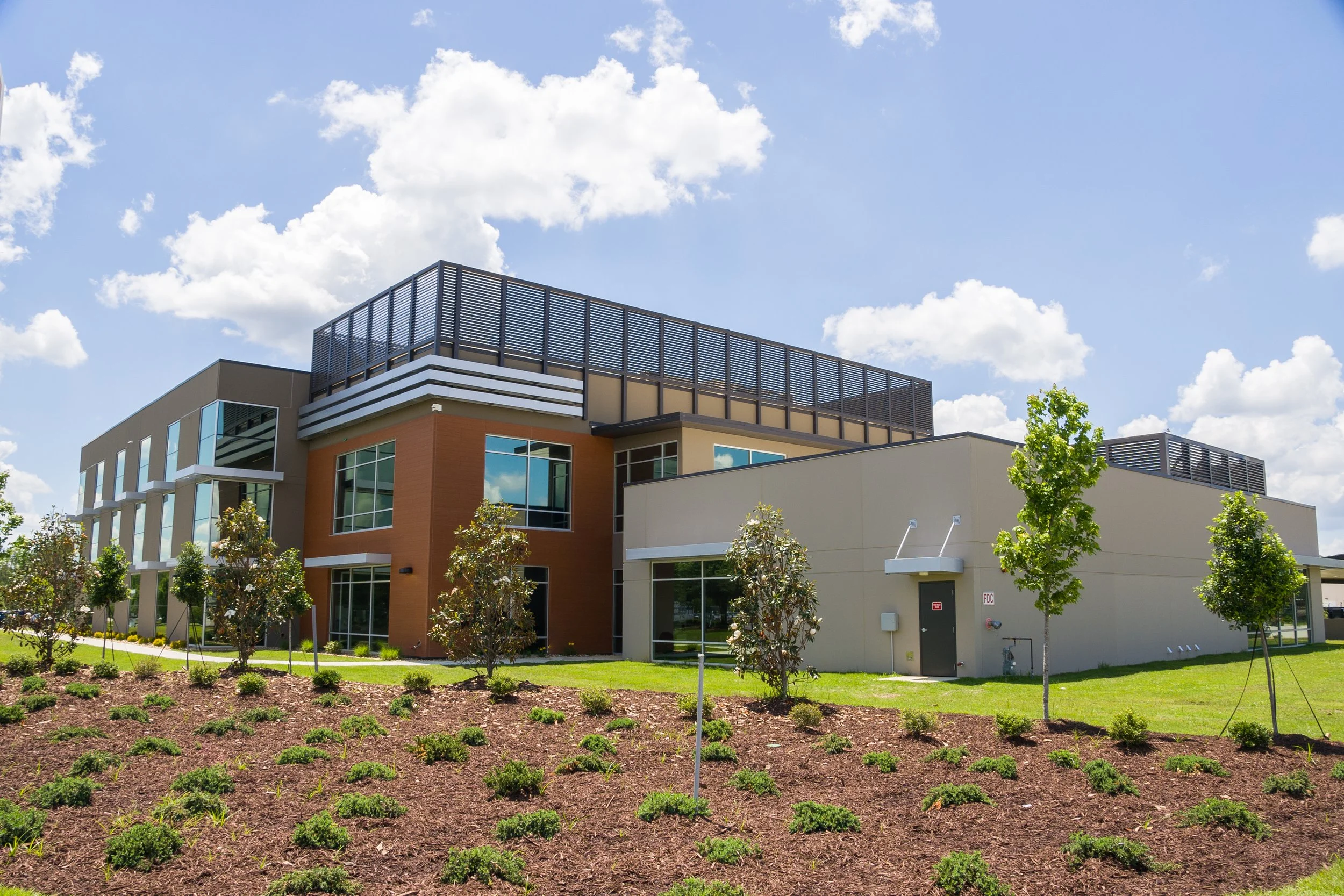 Modern multi-story building with landscaped garden and young trees, under a partly cloudy sky.