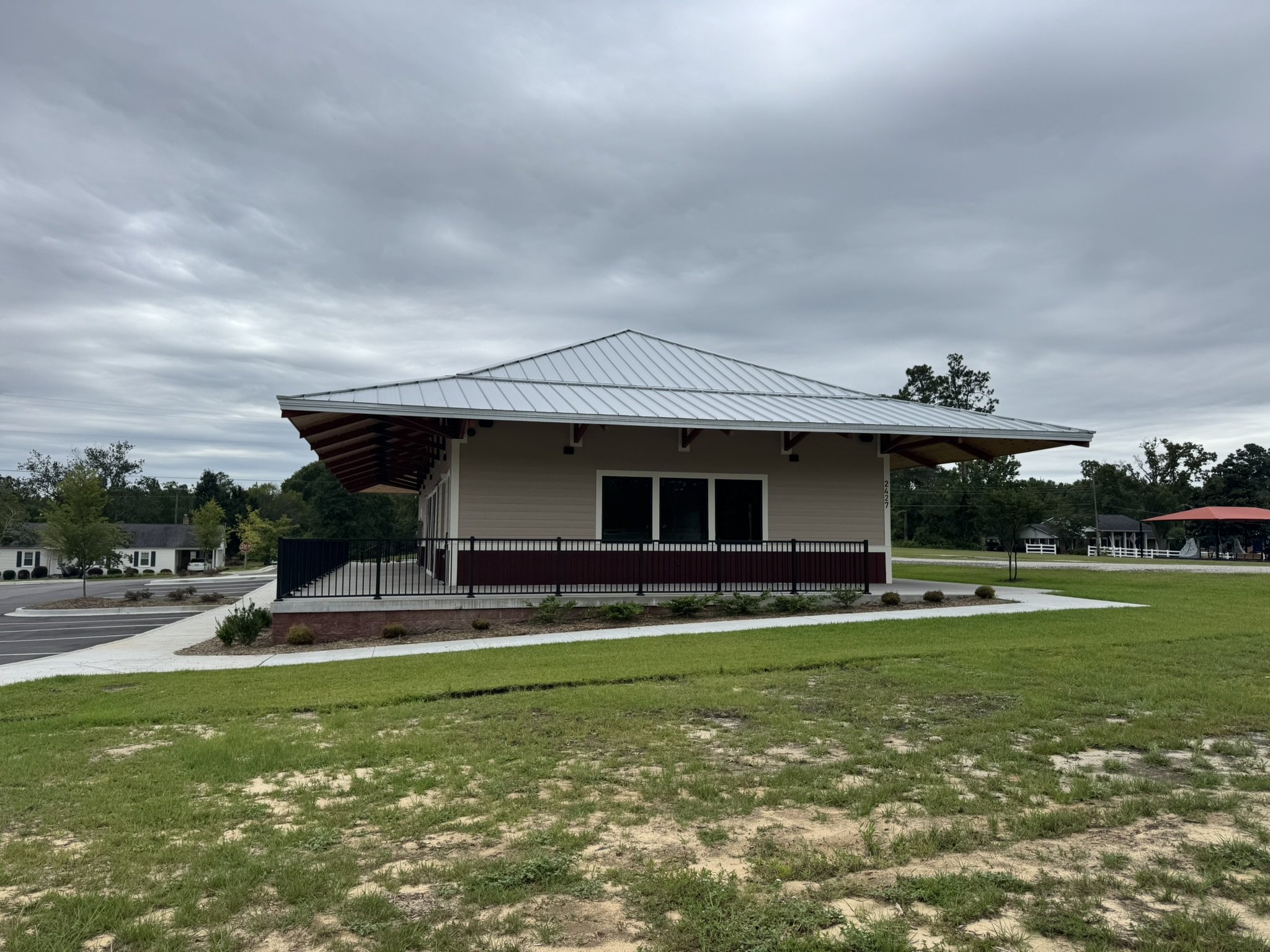 An outdoor scene with a modern house that has a grey metal roof, a small balcony, and large windows. The foreground features a grassy patch with some dirt patches, and the background includes trees and a cloudy sky.