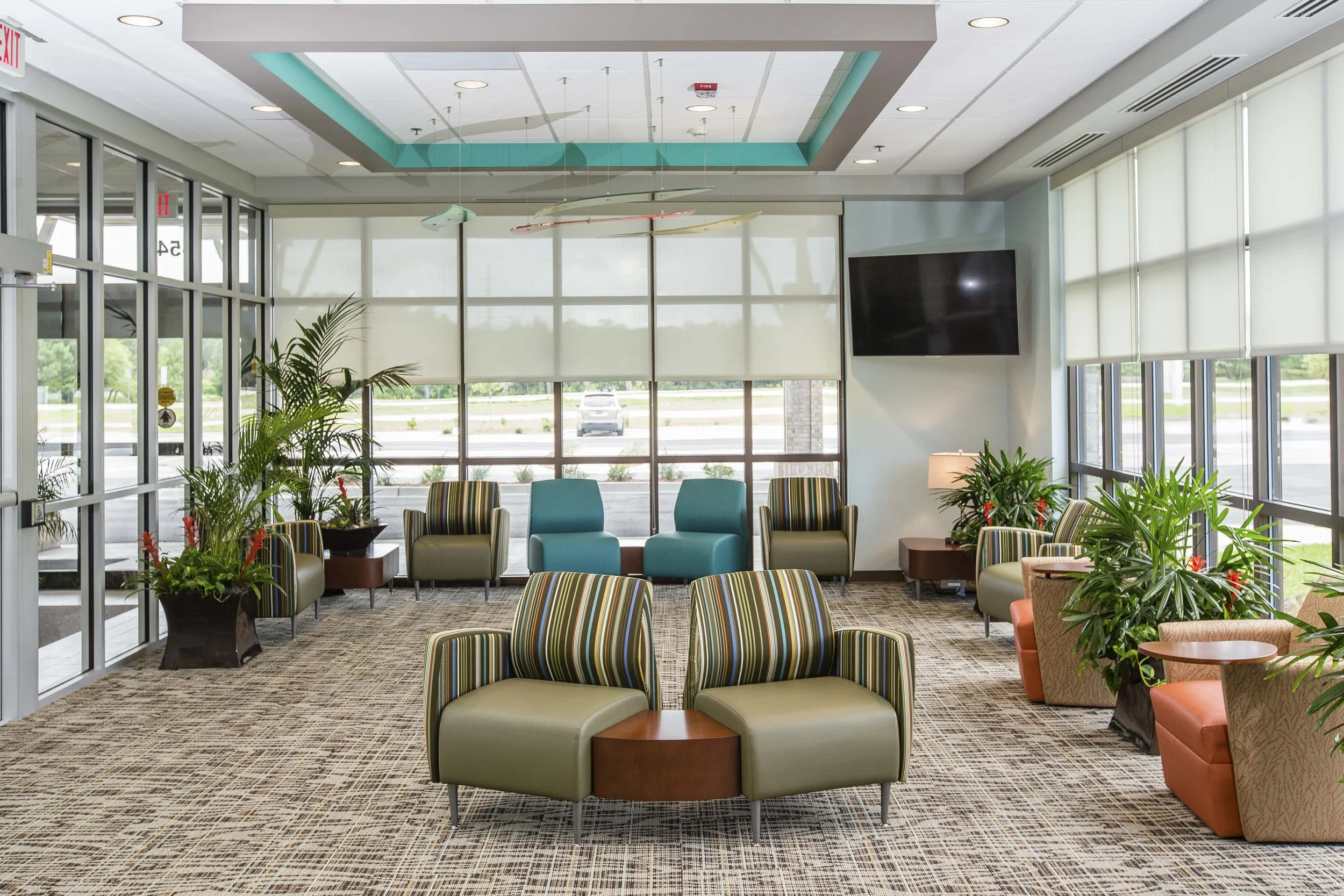 Interior of a modern waiting area with colorful striped and solid chairs, large windows with white blinds, indoor plants, a wall-mounted TV, and ceiling light fixtures.