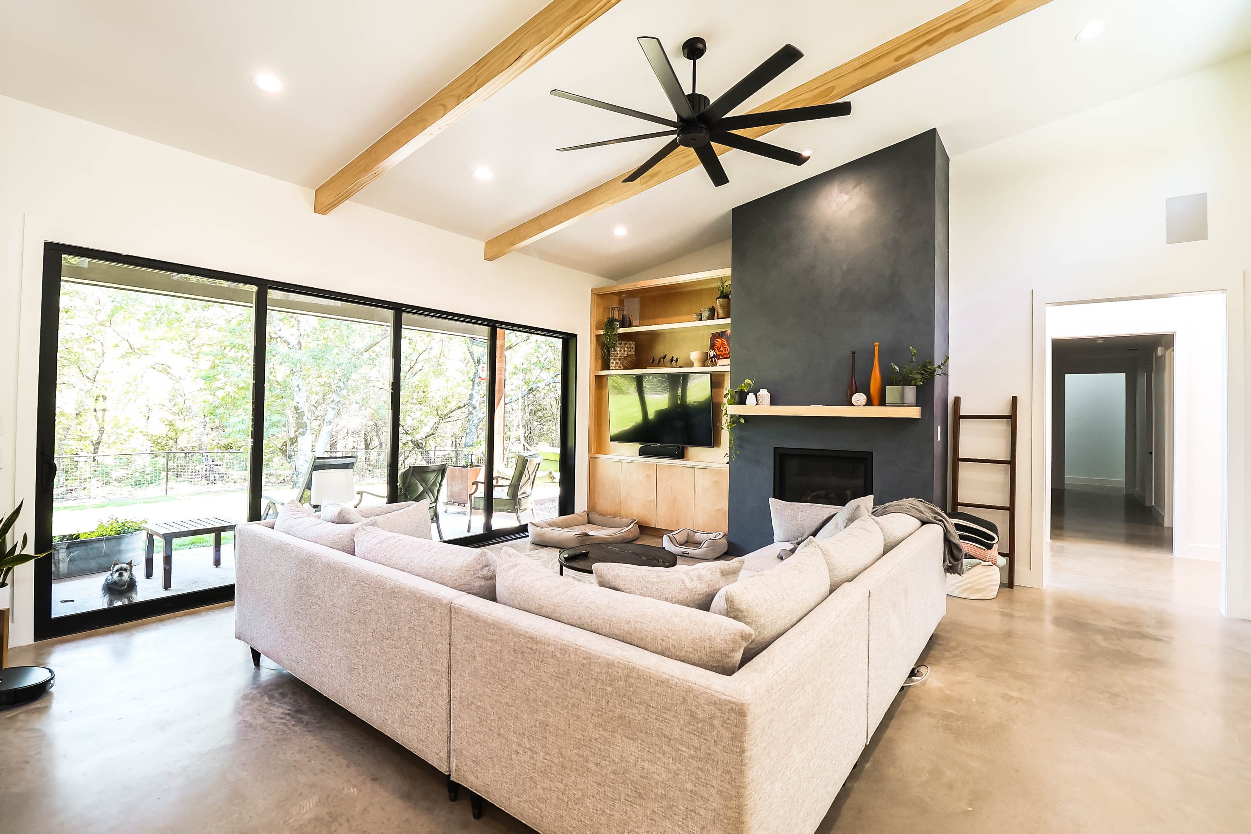 Living room with beige sectional sofa, large sliding glass door with a view of outdoor patio and trees, black ceiling fan, wooden beams, built-in shelves with decor, and a TV, with a dog at the sliding door.