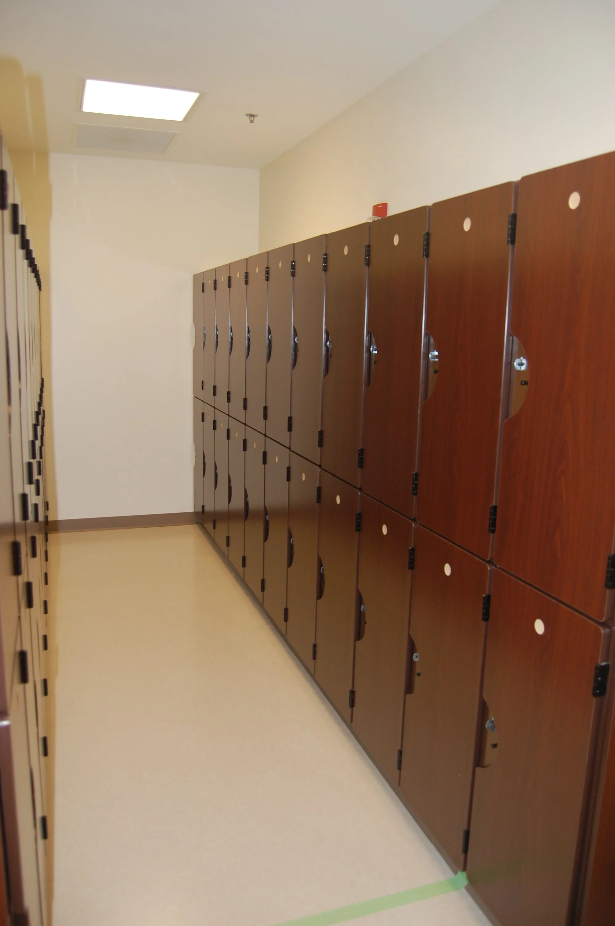 A hallway with a row of brown lockers on the right wall and a white floor with a green tape line near the bottom right corner.