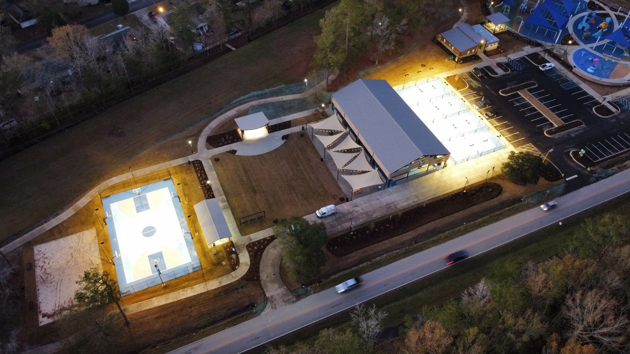 Aerial view of a park at night featuring a basketball court, a skate rink, a playground with slides, and a parking lot, all illuminated by lights.