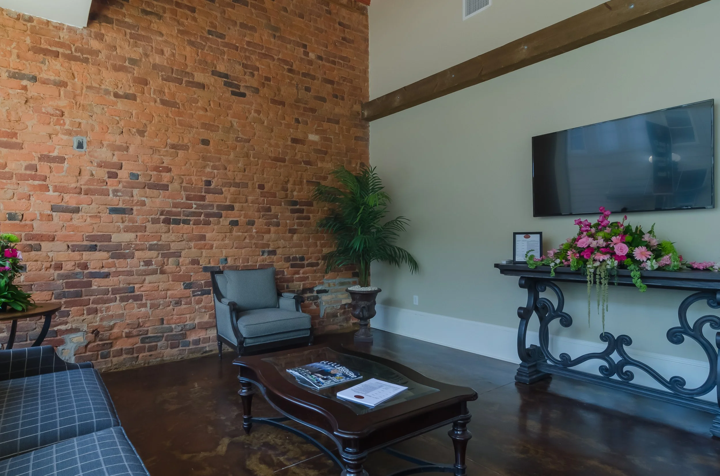 Interior room with exposed brick wall and beige wall, featuring a mounted flat-screen TV, a potted plant, a gray armchair, a dark coffee table with magazines and papers, and a black table with a floral arrangement of pink and purple flowers.
