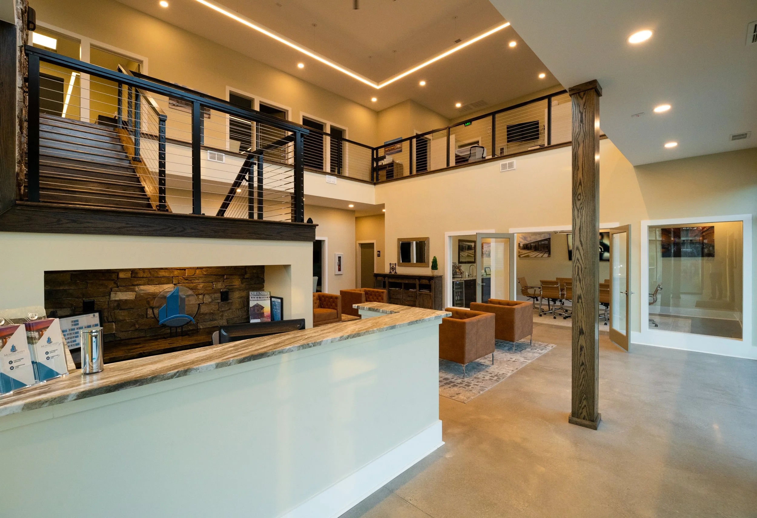 Interior of a modern office or hotel lobby with a reception desk, seating area with orange chairs, an upper-level walkway with black railings, and ceiling lighting.
