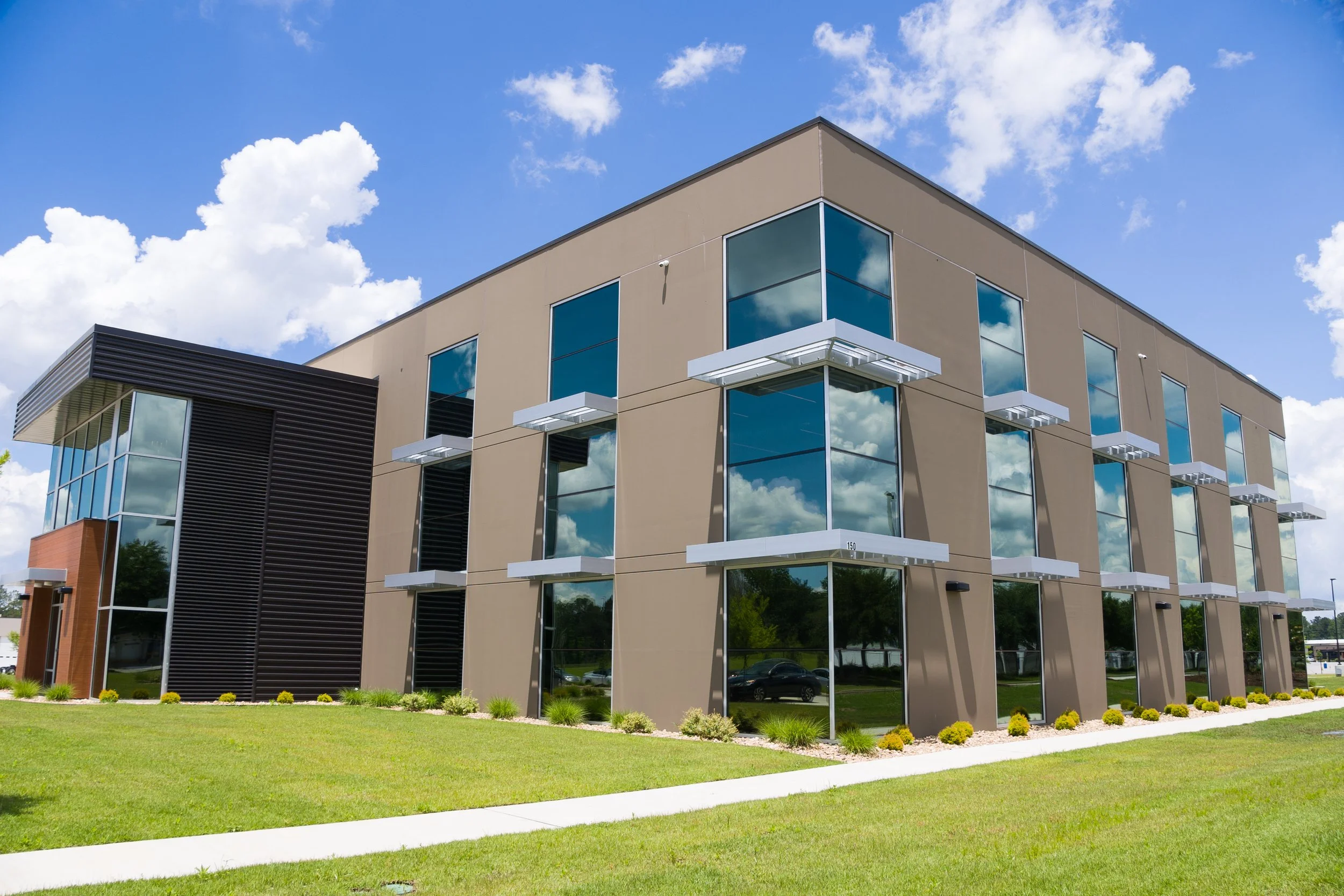 Modern multi-story building with large reflective glass windows and beige exterior walls. The building has white balcony sunshades and a landscaped lawn with small bushes in the foreground. The sky is partly cloudy with white clouds and blue sky.